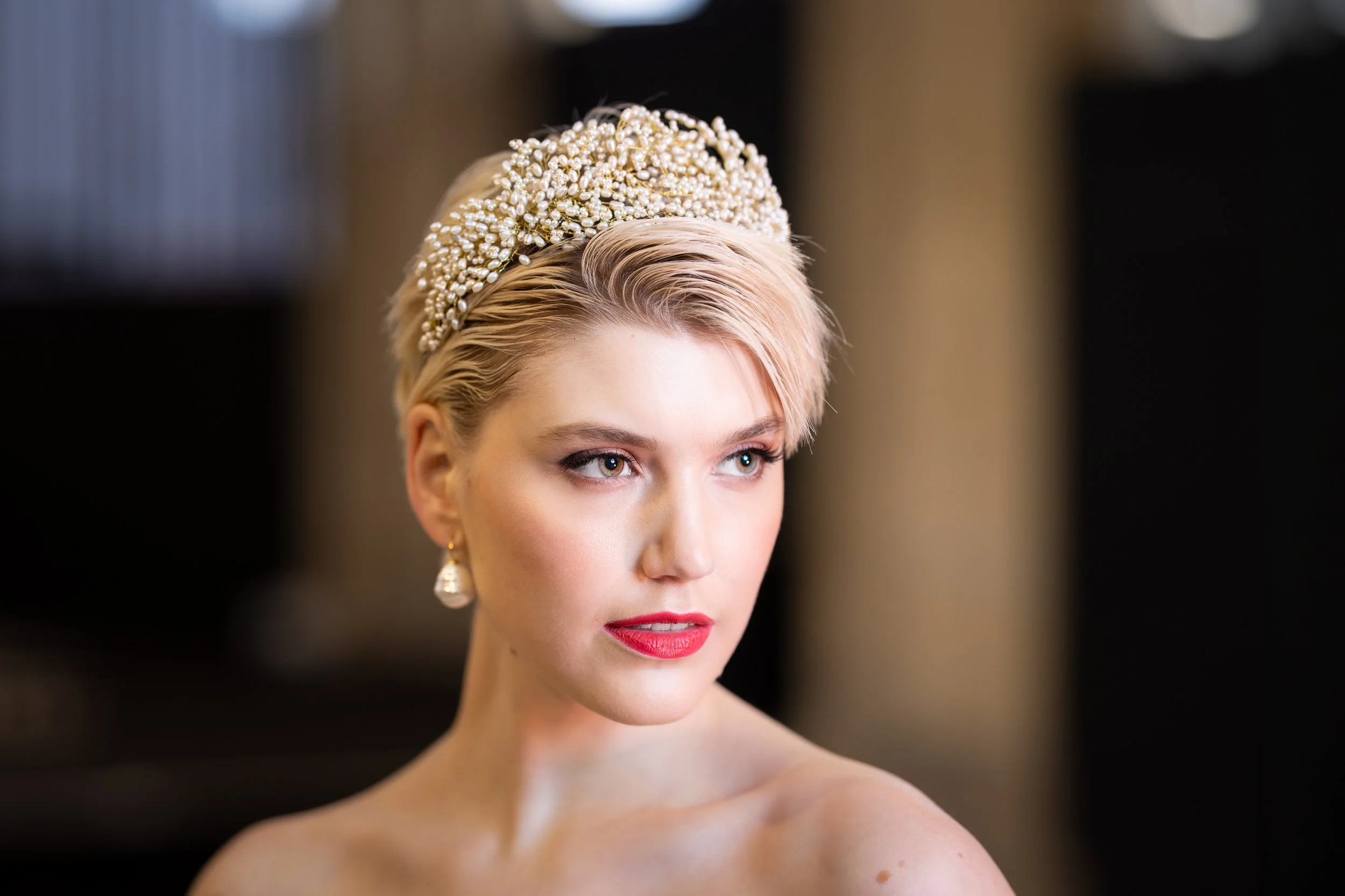 Portrait of a blonde woman with styled hair and elegant jewelry, wearing pearl earrings and a decorative pearl-studded headband, with a blurred background.