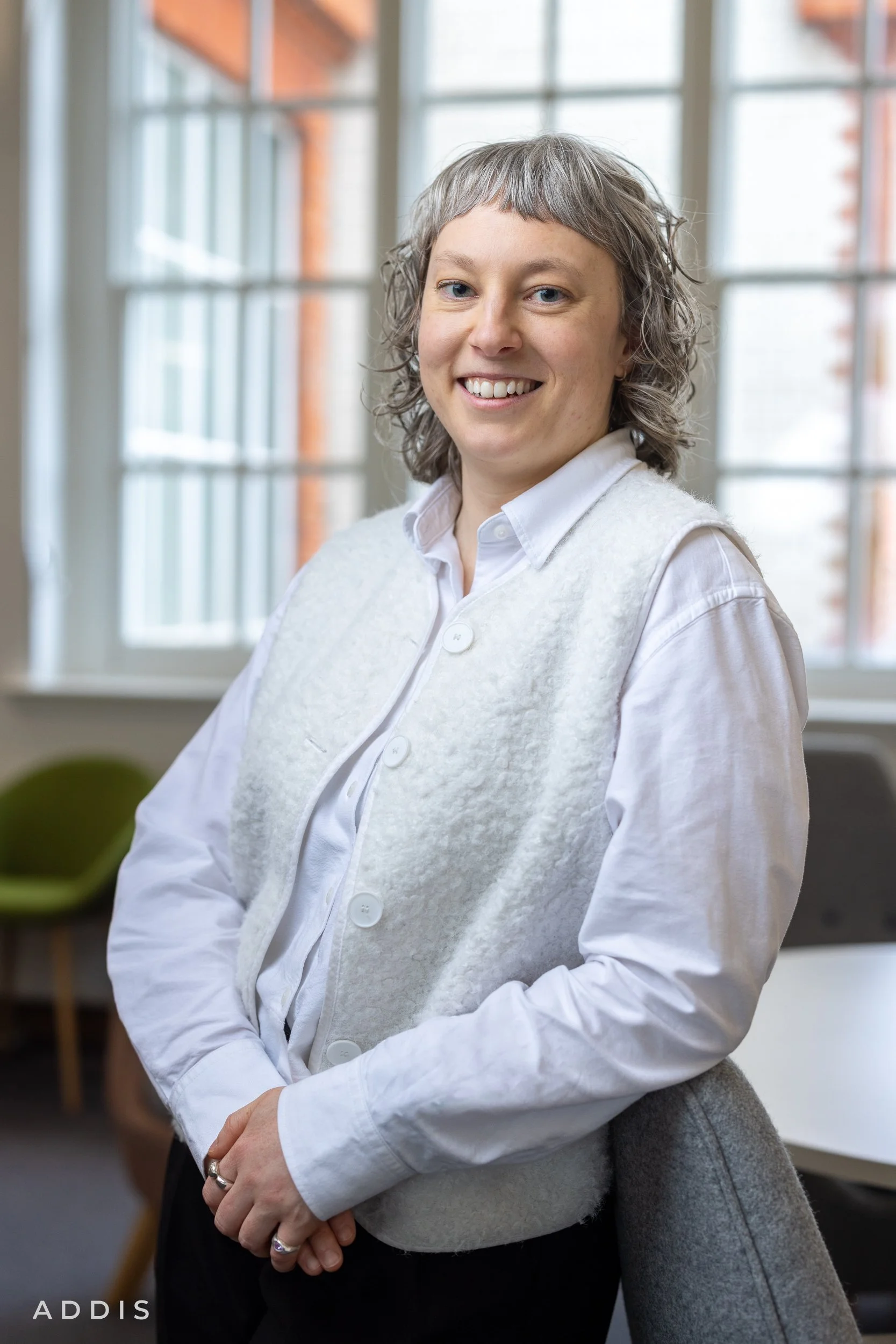 A woman with short curly gray hair and blue eyes, wearing a white button-up shirt and a gray vest, smiling in front of large windows with brick and white framing.