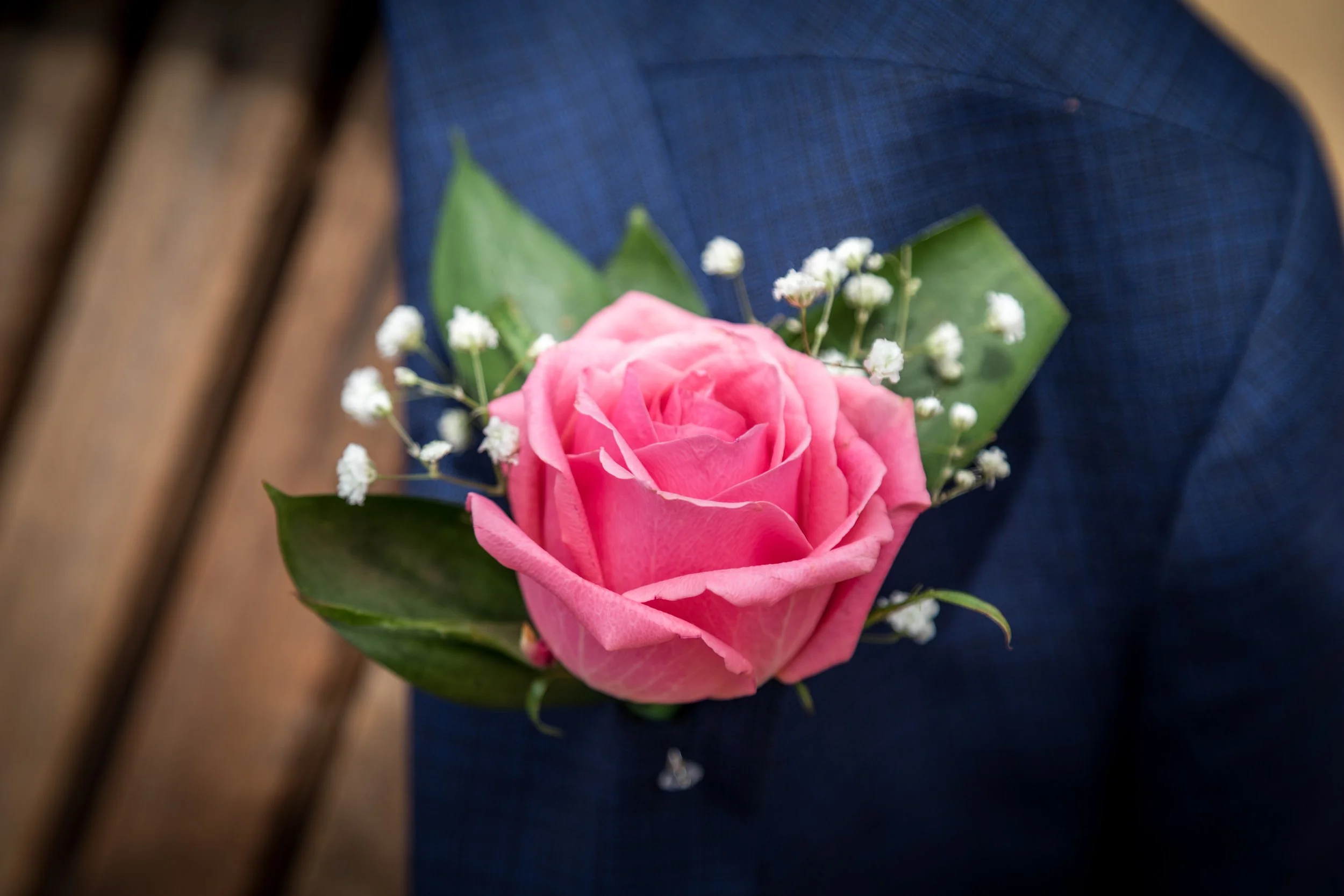 Close-up of a pink rose boutonniere with small white filler flowers and green leaves, pinned to a person's navy blue suit.