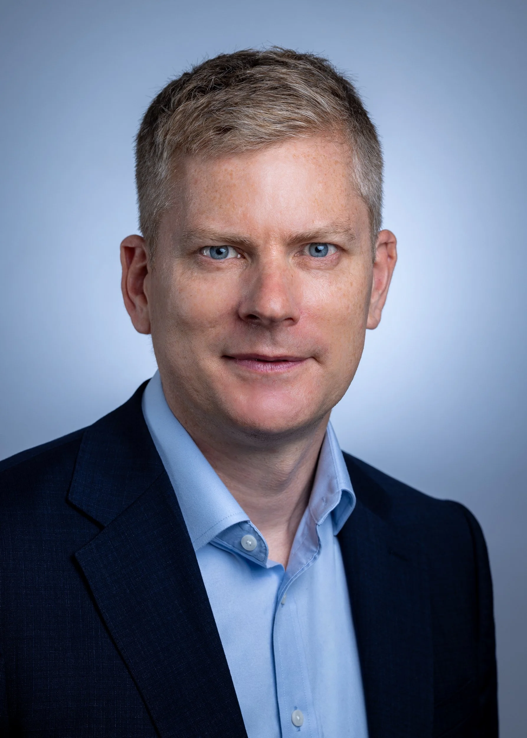A portrait photo of a man with short light brown hair, blue eyes, wearing a dark suit jacket and a light blue collared shirt, against a plain light blue background.