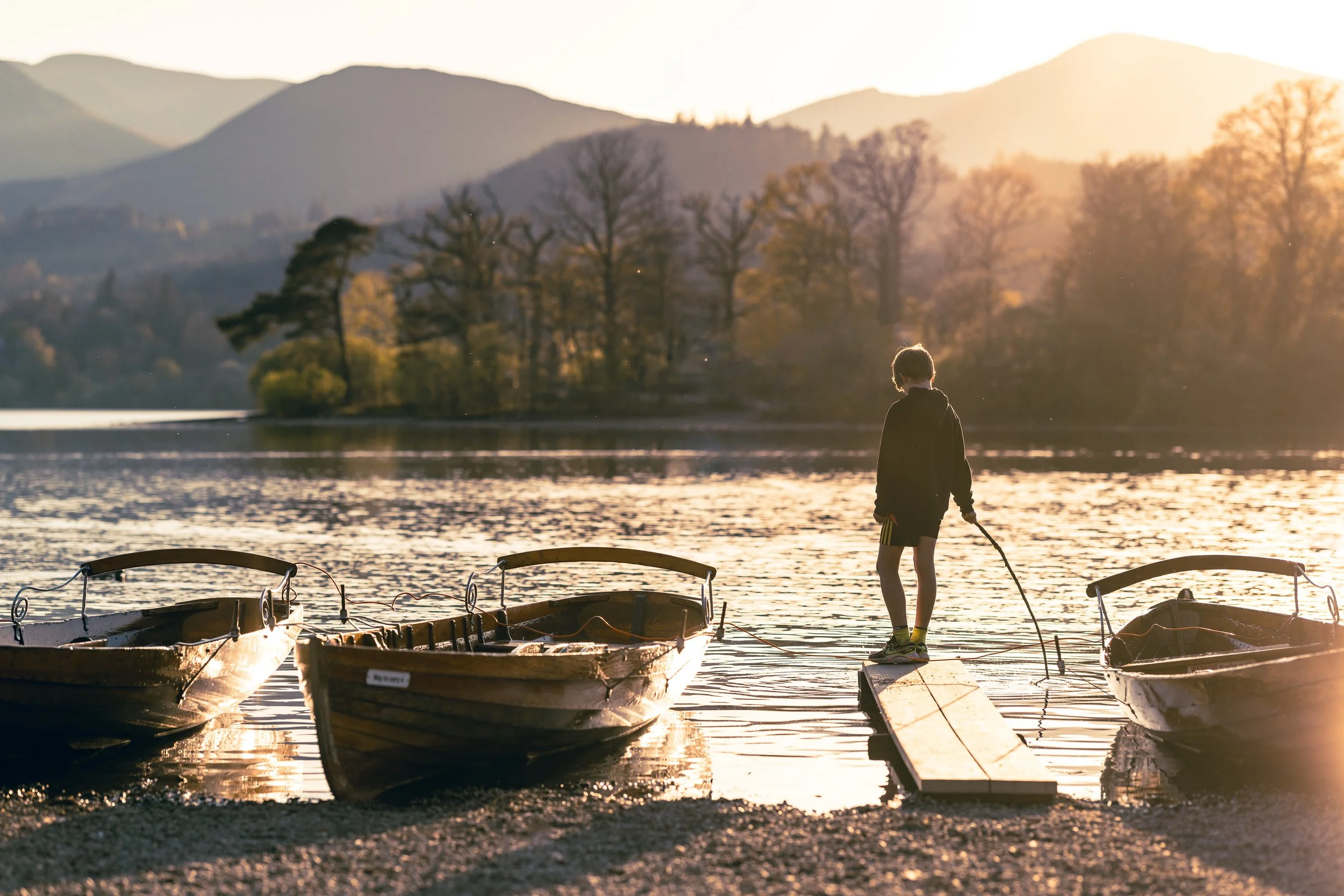 A person standing on a dock by the water during sunset, surrounded by boats and mountains in the background.