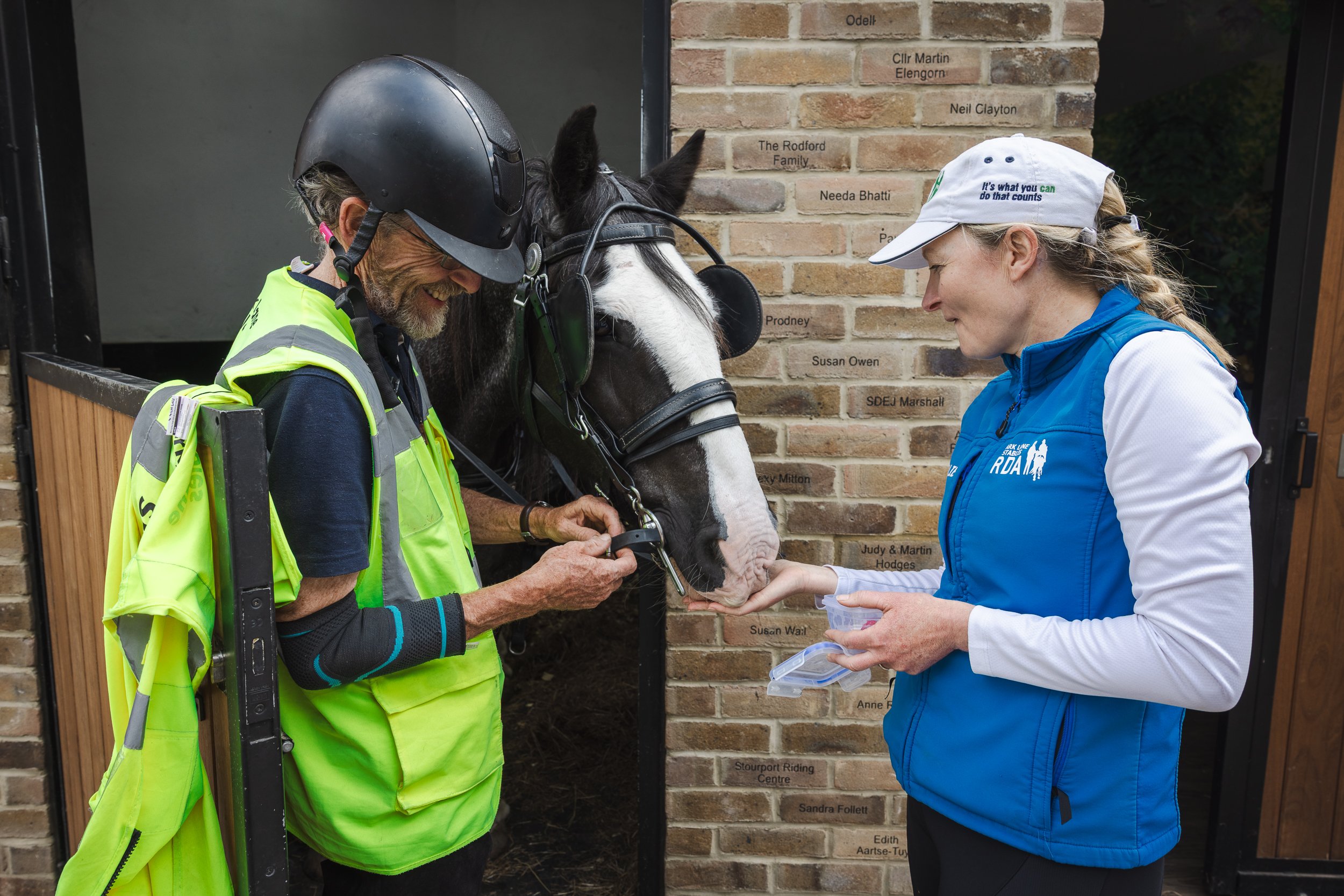 A Park Lane Stables horse and someone in a black helmet and yellow safety vest interacts with the horse, which wears a bridle. Natalie O'Rouke in a white cap and blue vest looks at the horse. The brick wall behind them has engraved names.
