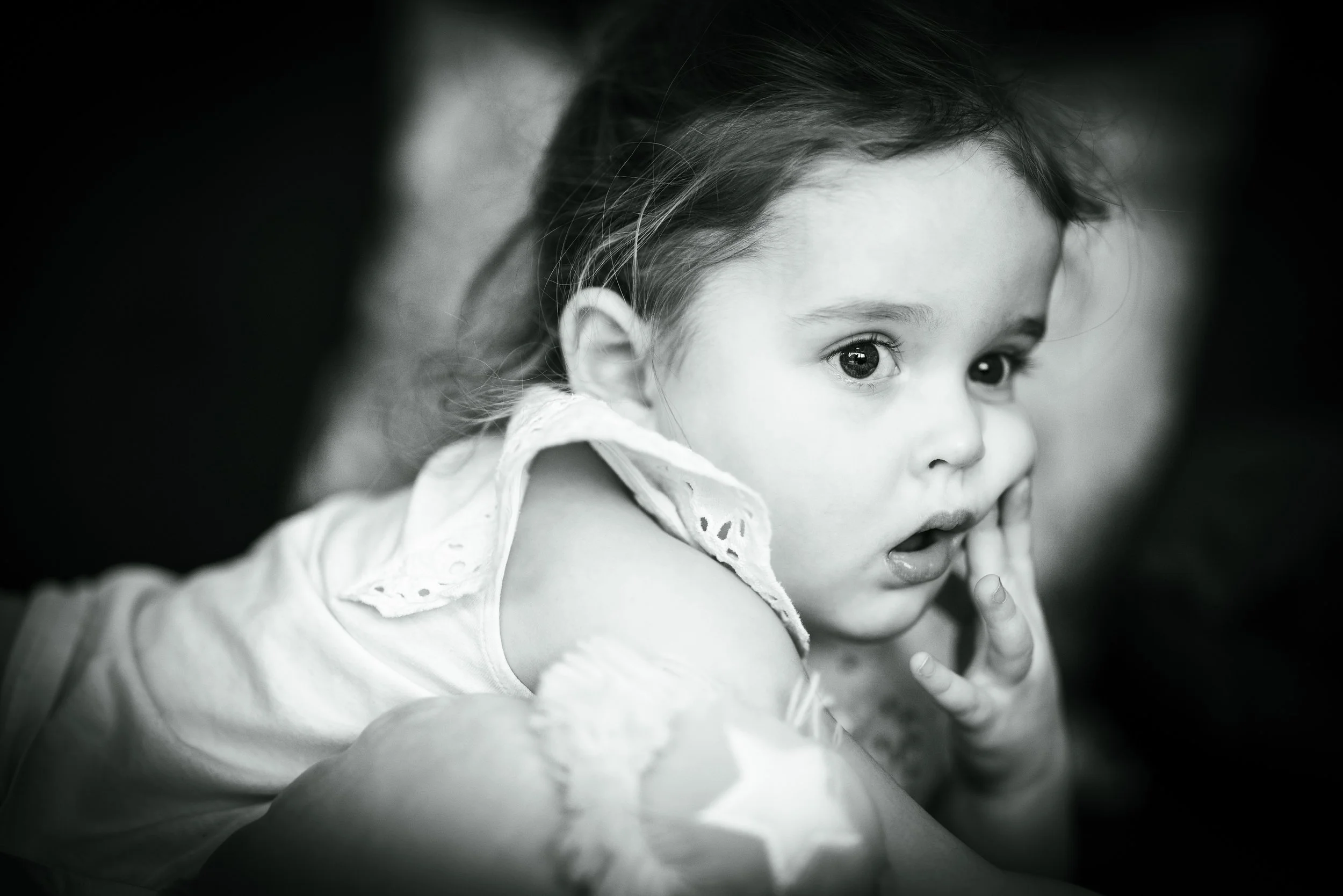 A black and white photo of a young girl with big eyes and dark hair, wearing a light-colored shirt with eyelet details on the collar, looking directly at the camera with her finger near her mouth.