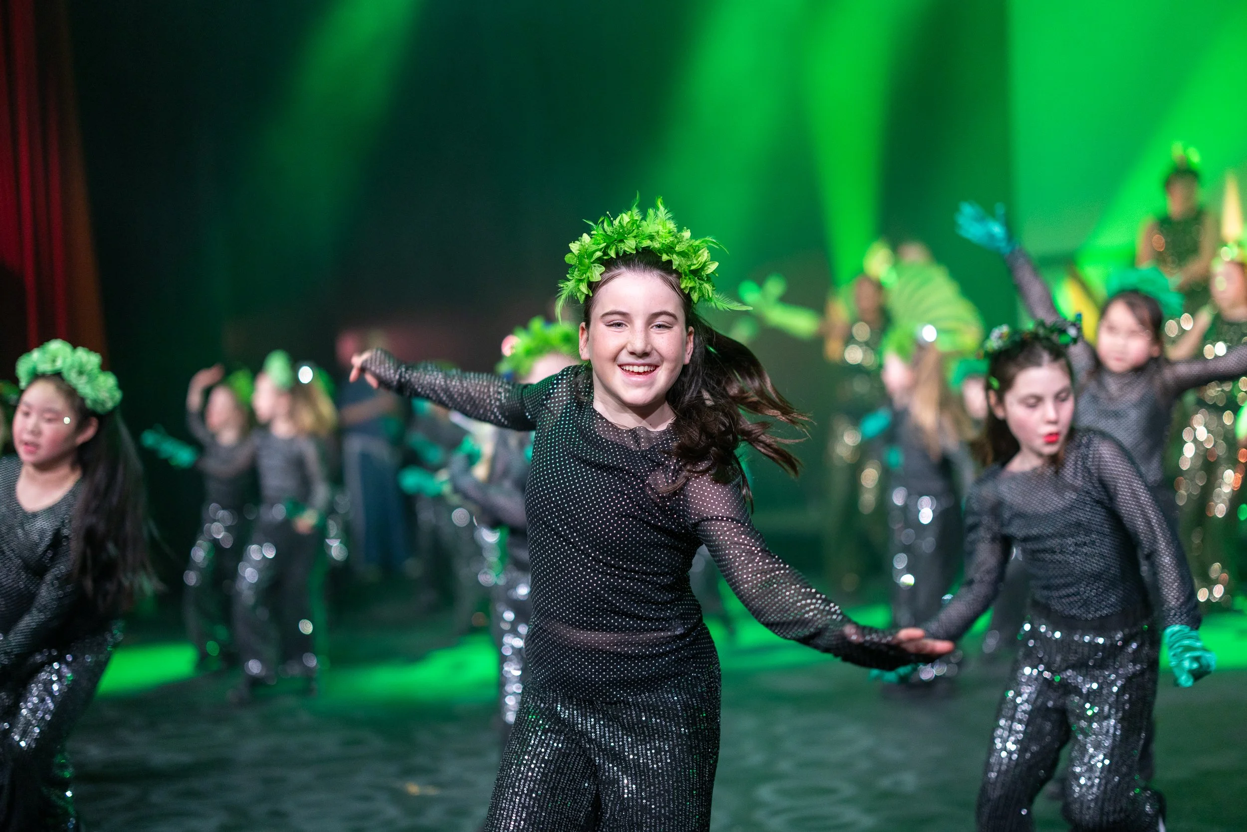Young dancers wearing black polka dot costumes with green leafy headpieces dancing on stage at Questors Theatre in Ealing with green lighting.