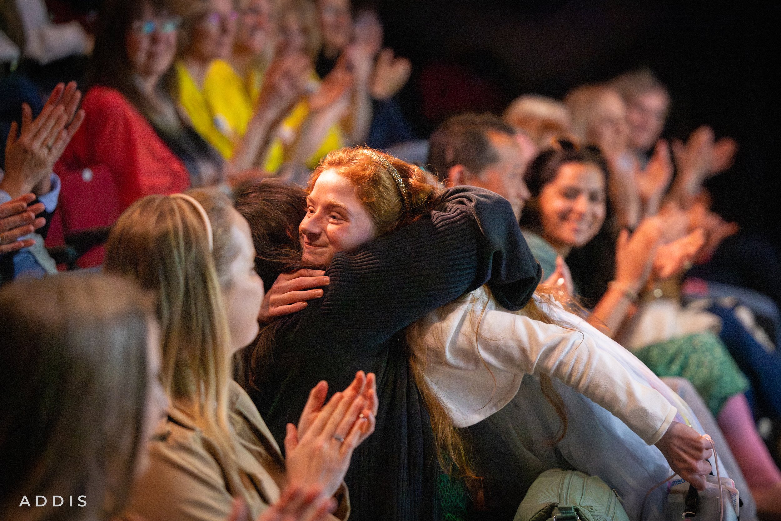A group of people sitting in an audience clapping, with a woman with red hair hugging another person in the foreground, smiling.