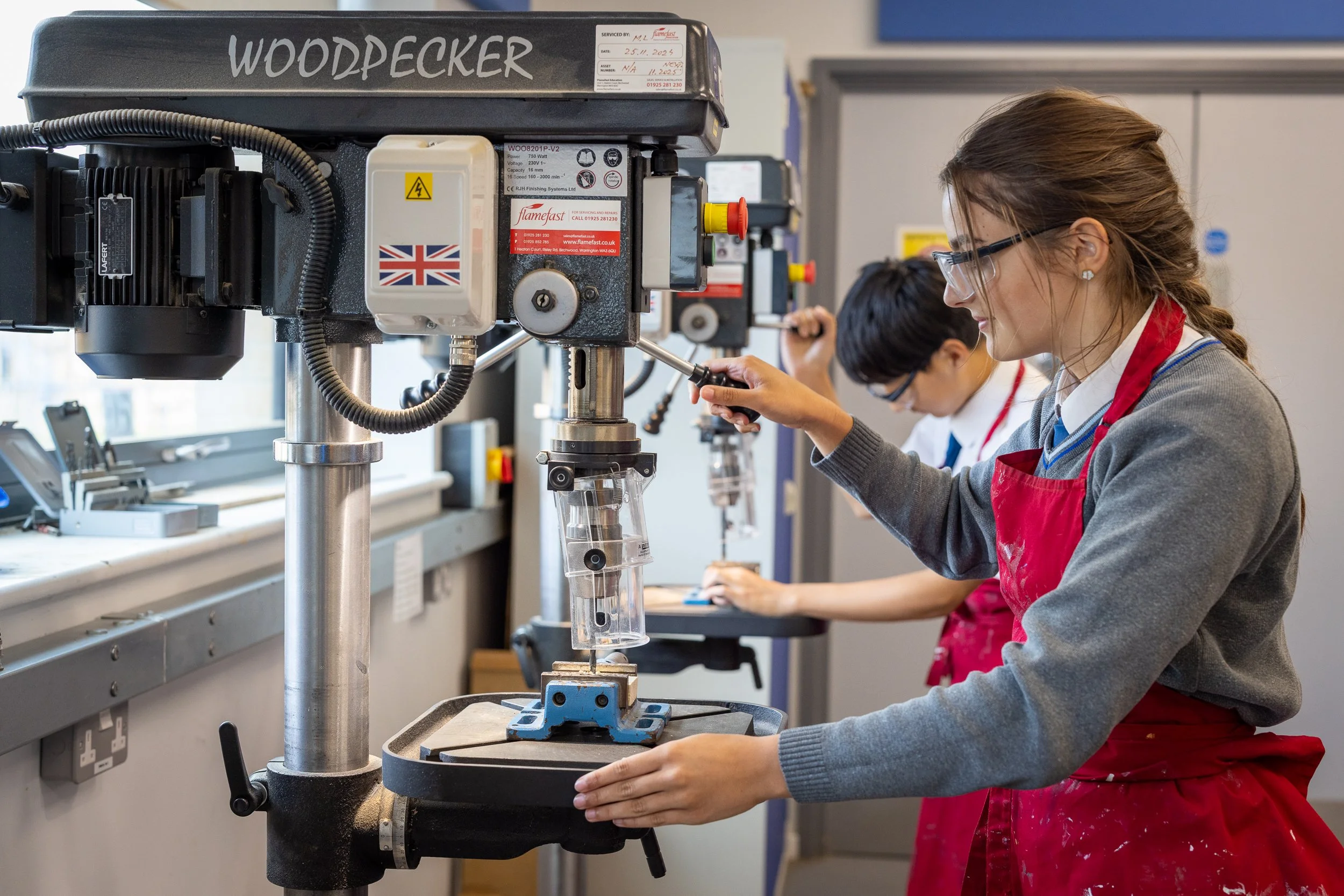 Two students in a workshop at The Matthew Arnold School using a milling machine, with the girl in the foreground adjusting the machine and wearing a red apron and glasses.