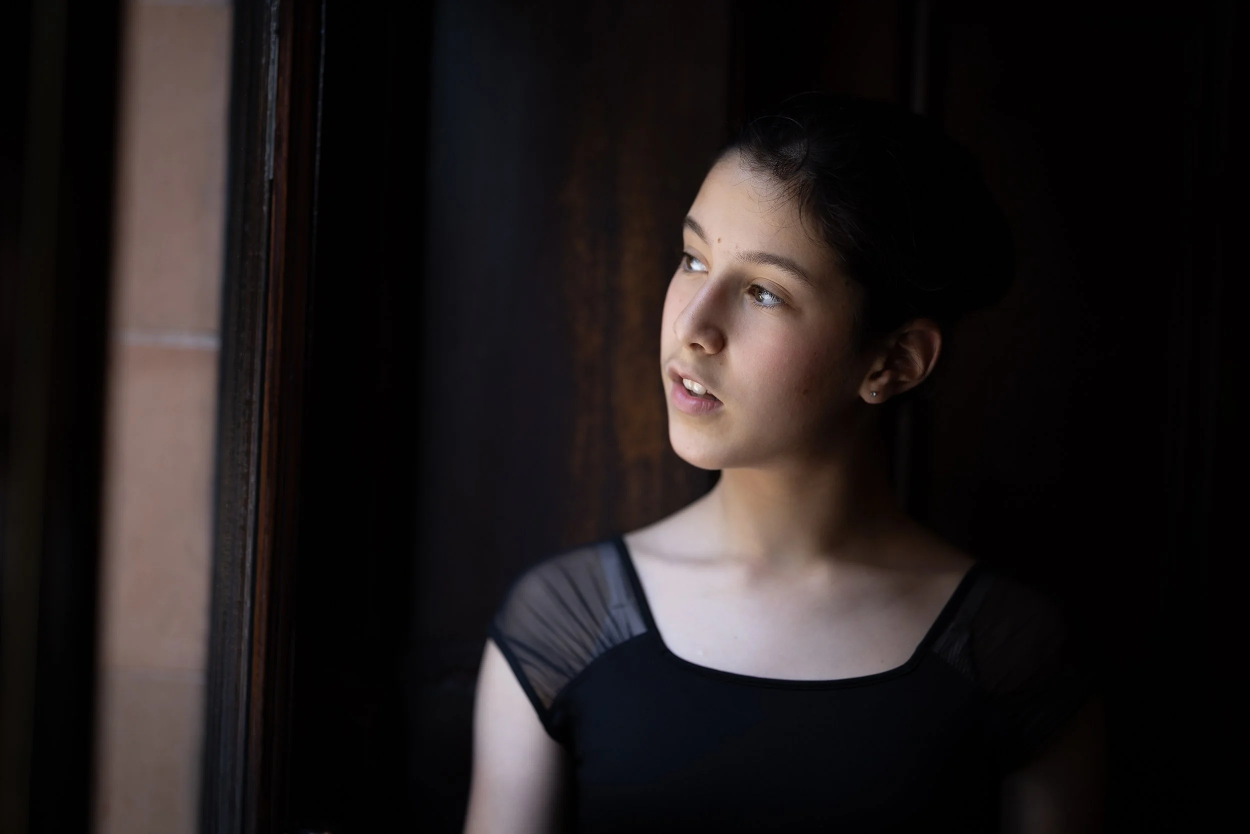 A young woman with dark hair styled back is looking out of a window with soft natural light illuminating her face.