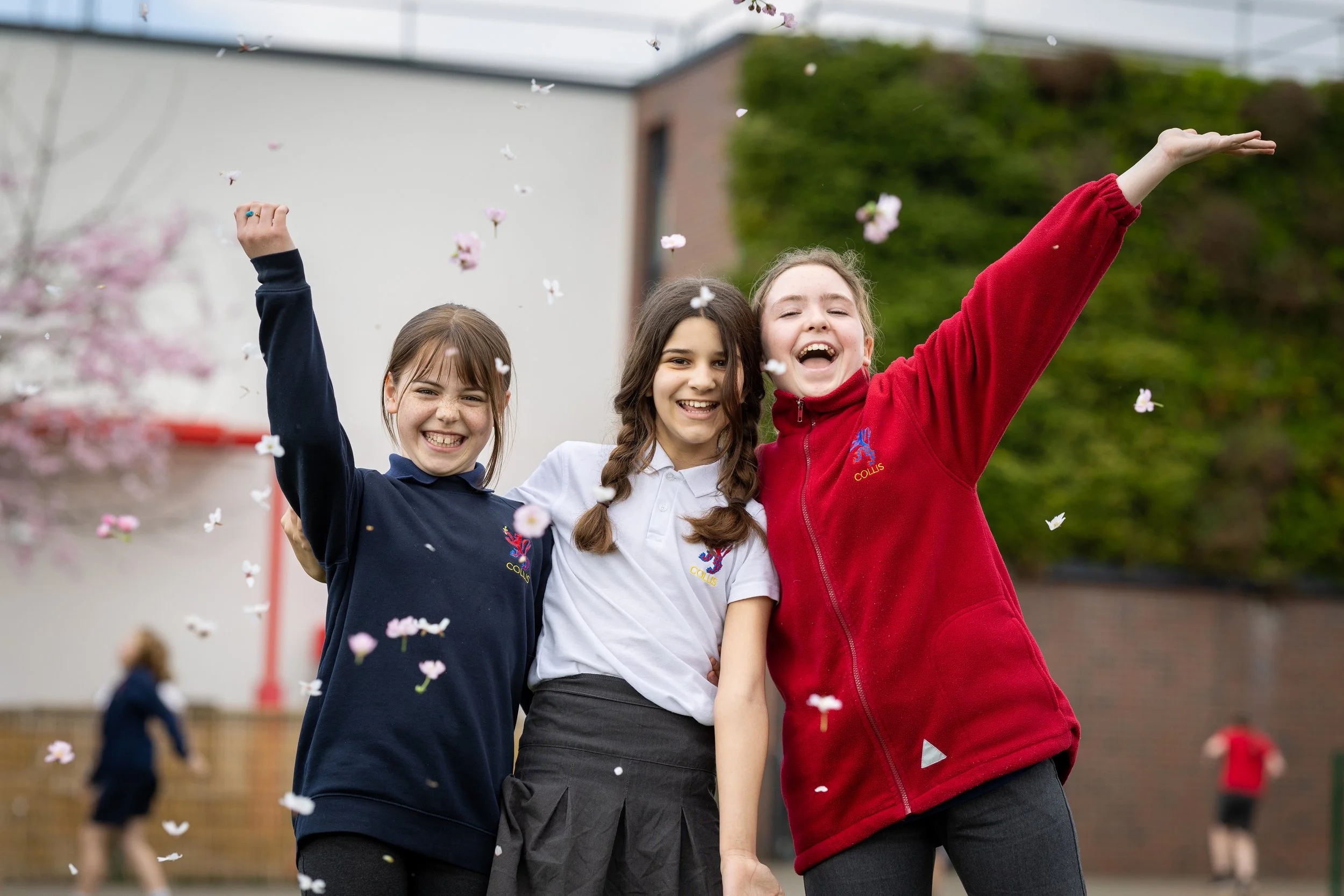 Three smiling young girls, wearing school uniforms from Colis, are outdoors during the day, playing and throwing flower petals in celebration.