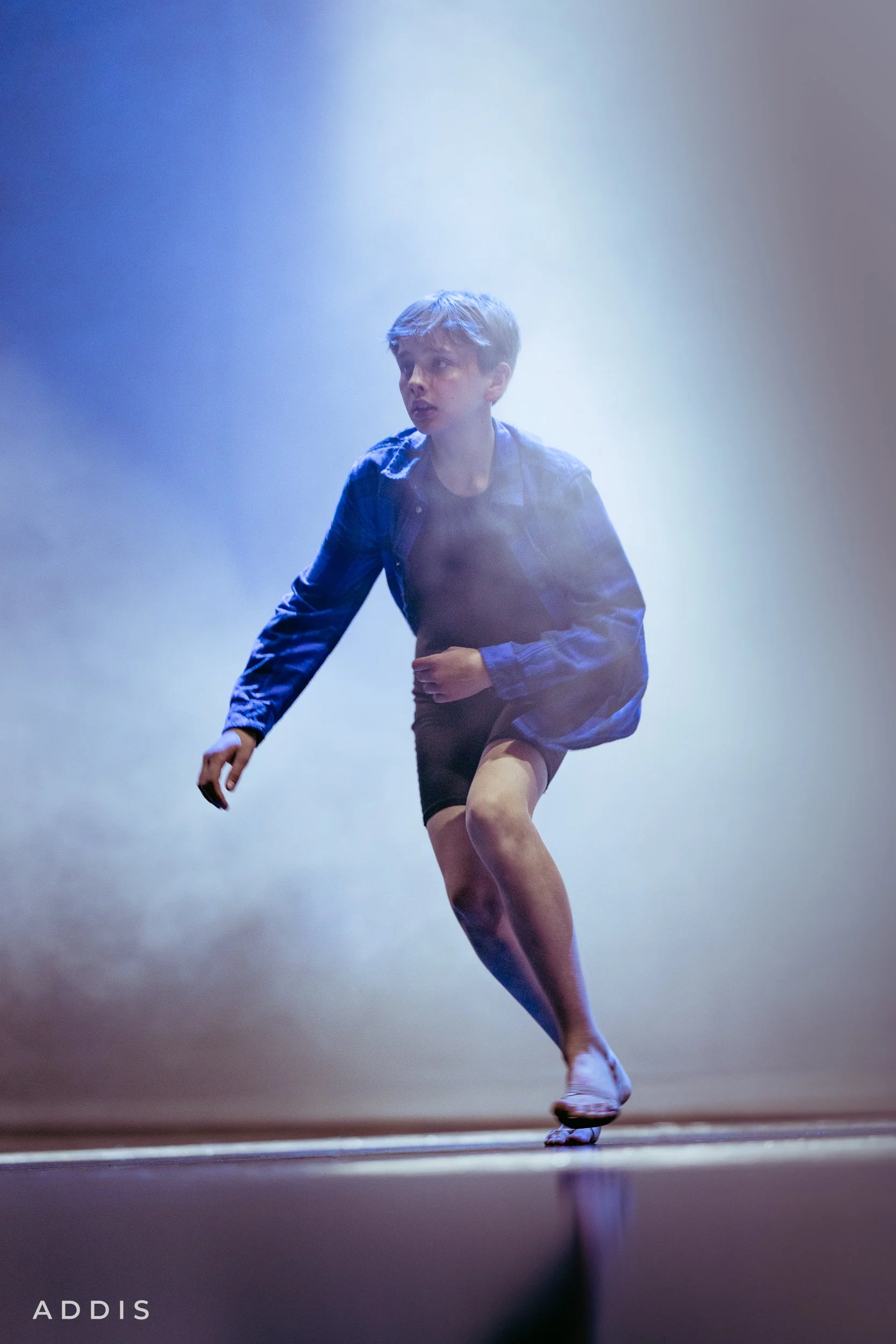 A young male dancer in a blue jacket and black shorts performing on stage with a smoky background.
