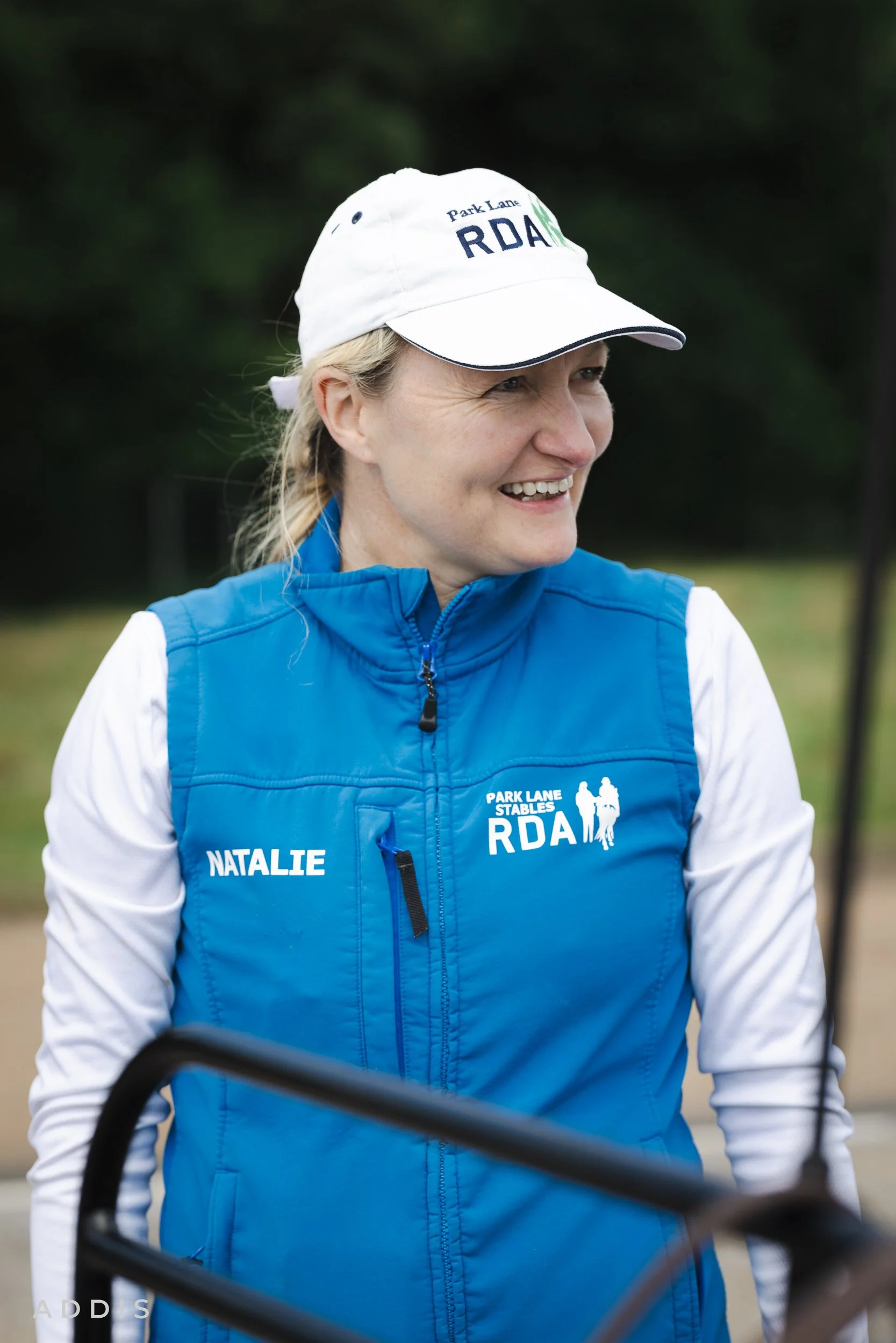 Natalie O'Rouke smiling outdoors wearing a white cap with green and black writing, a blue vest with white sleeves and text, and a zipper, with a background of dark green trees.