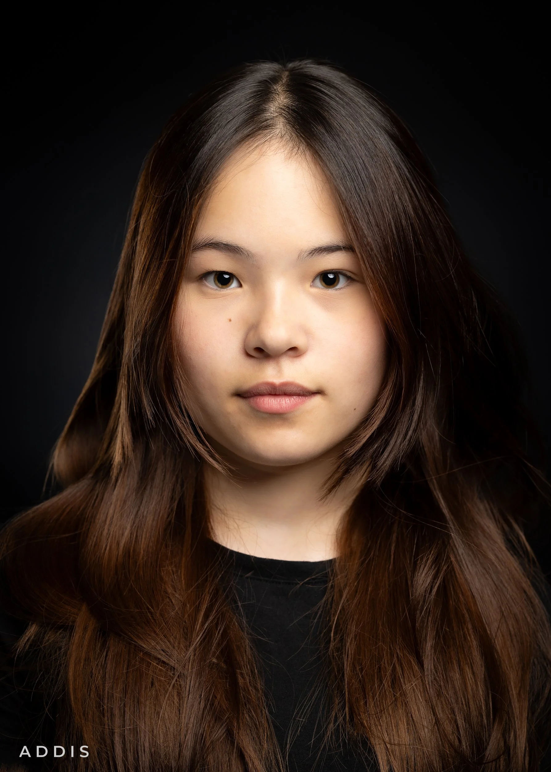 Close-up portrait of a young woman with long, brown hair wearing a black top against a black background.