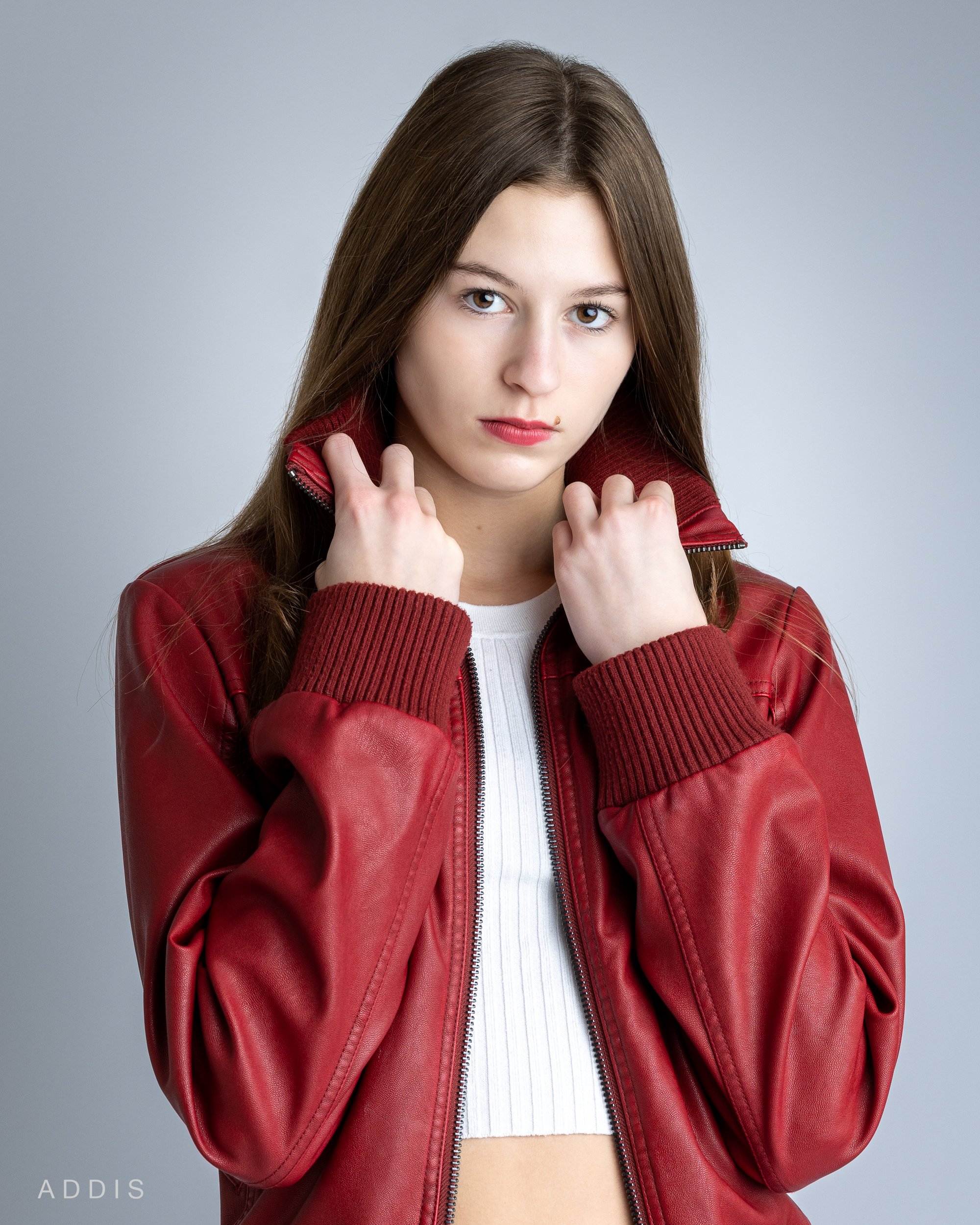 A young woman with long brown hair wearing a red leather jacket and a white crop top, facing forward with her hands holding the collar of her jacket, against a light gray background.