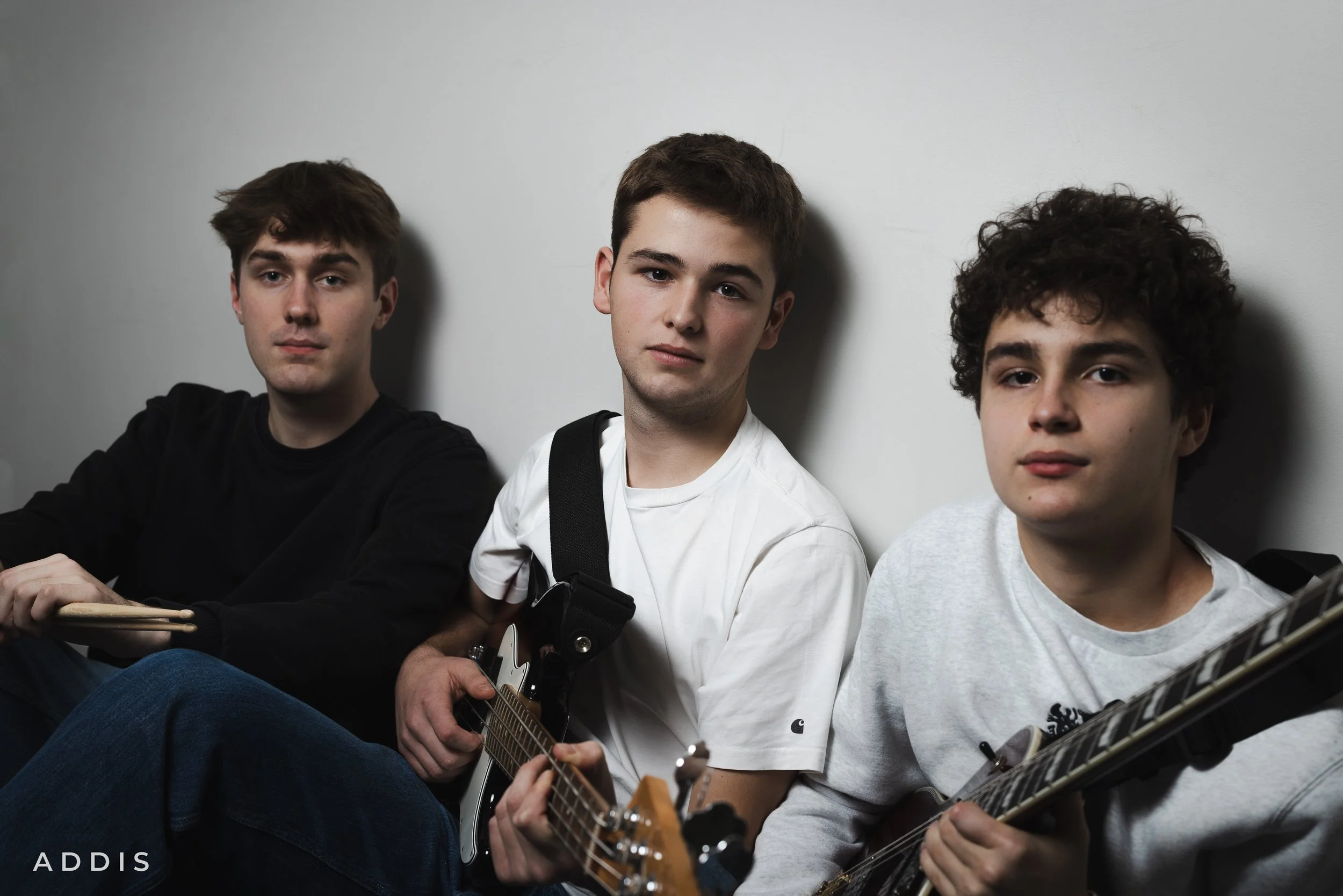 Three young men sitting against a white wall, each holding a guitar, looking at the camera.