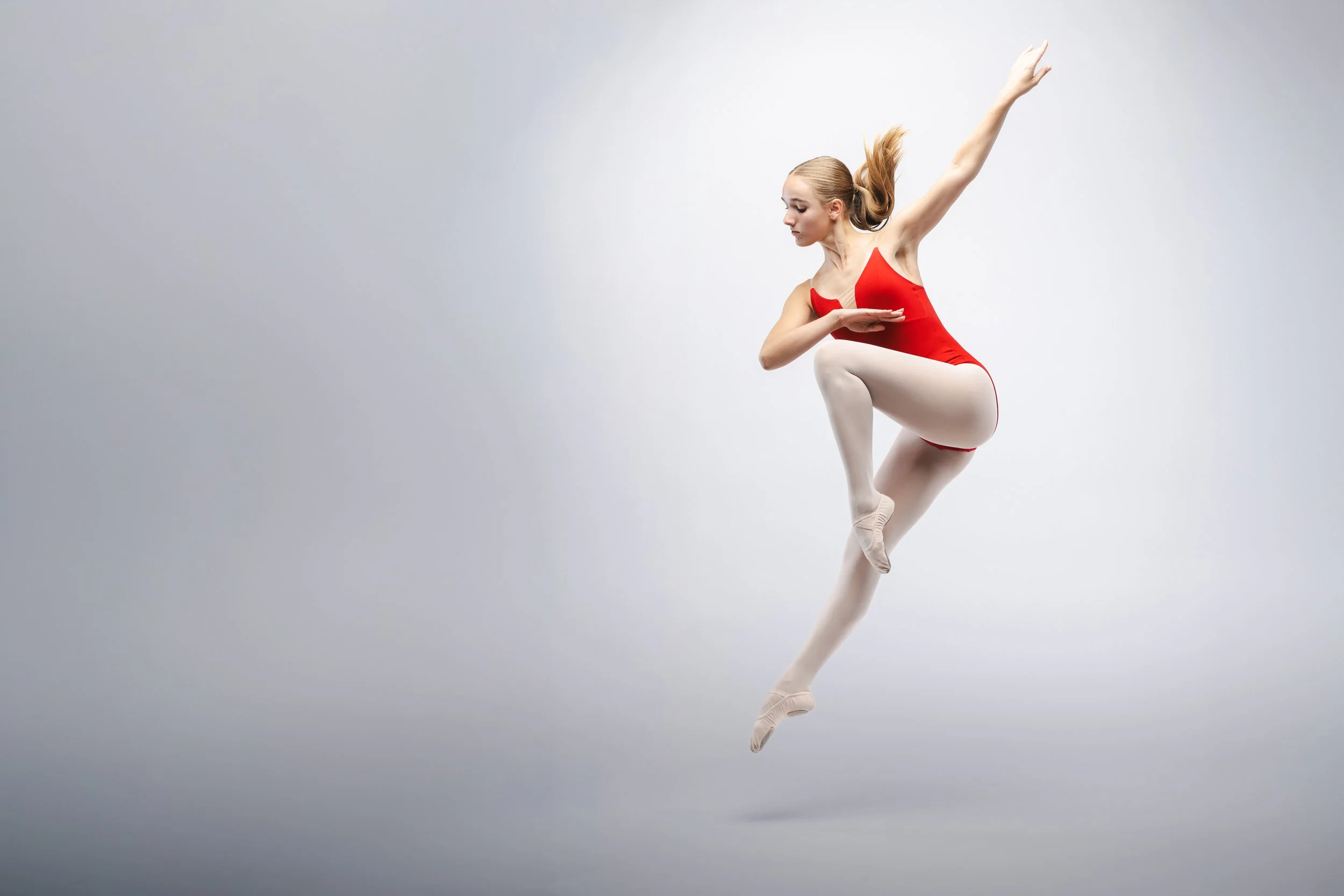 A ballet dancer in a red leotard and white tights performing a pose with one arm raised and the other arm at her waist, standing on one leg with the other leg bent and foot pointed.
