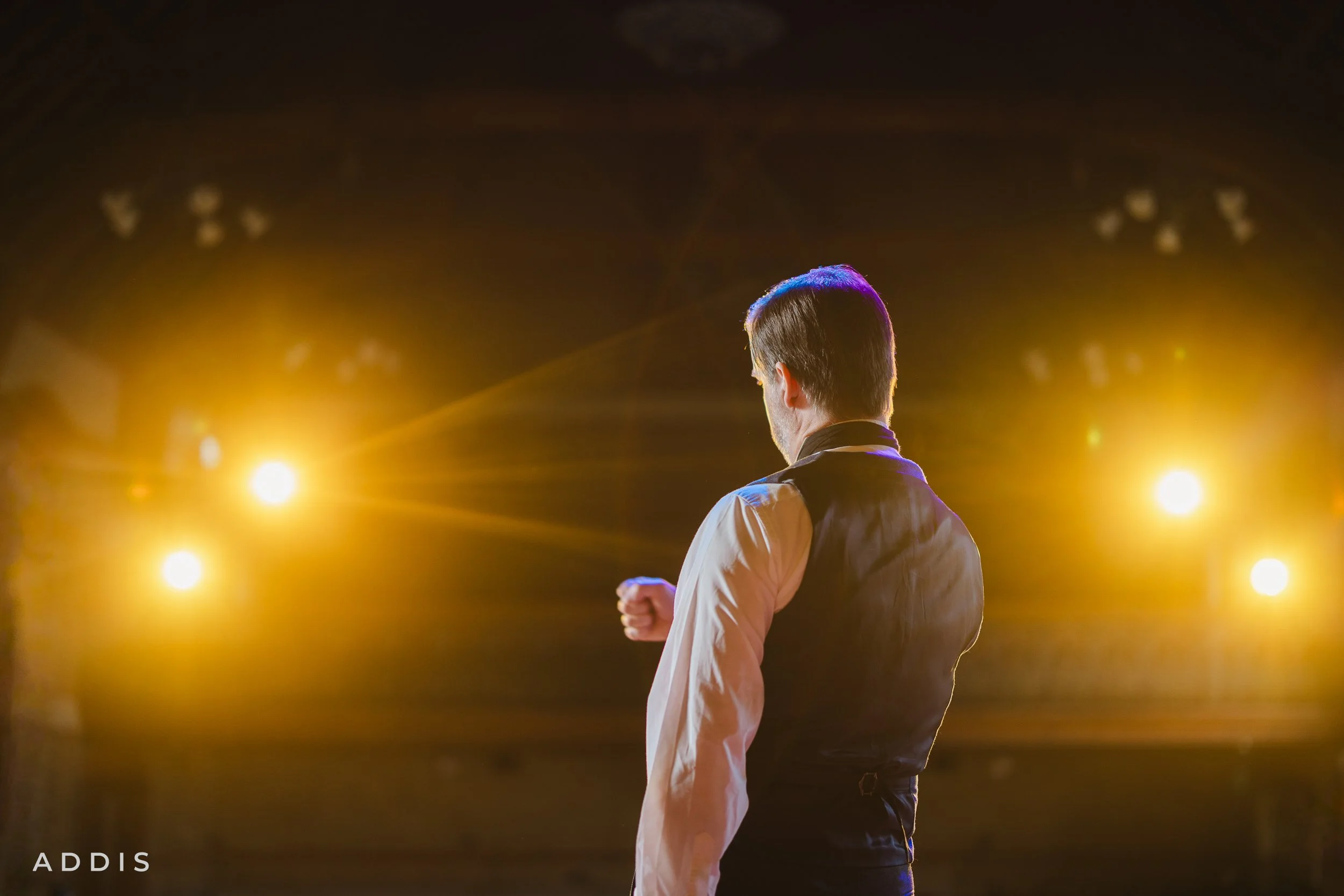 A man standing on stage in front of bright yellow stage lights, facing away from the camera with his right fist clenched.