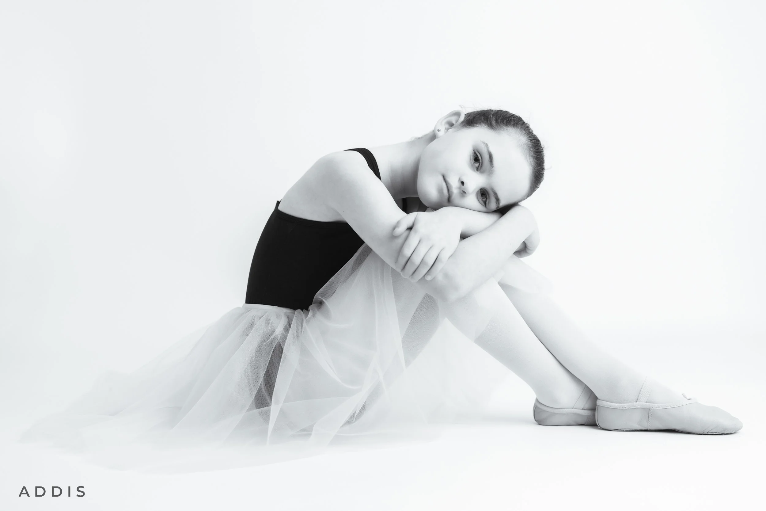 Black and white photo of a ballet dancer sitting on the floor, wearing a black leotard, a tulle skirt, and ballet shoes, with her arms wrapped around her knees, looking at the camera. The background is plain white.