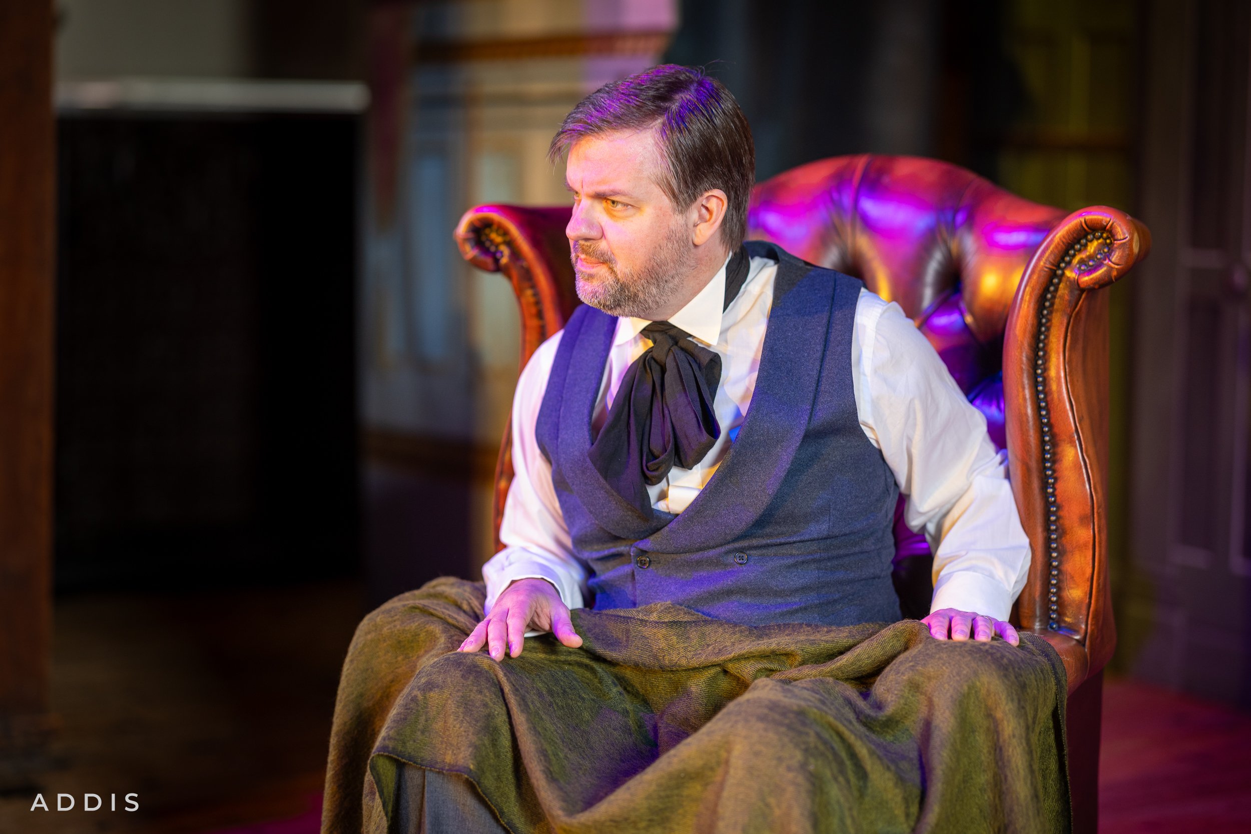 A man sitting on a vintage leather armchair, wearing period costume with a vest, shirt, and cravat, looking contemplative in a dimly lit room with bookshelves in the background.