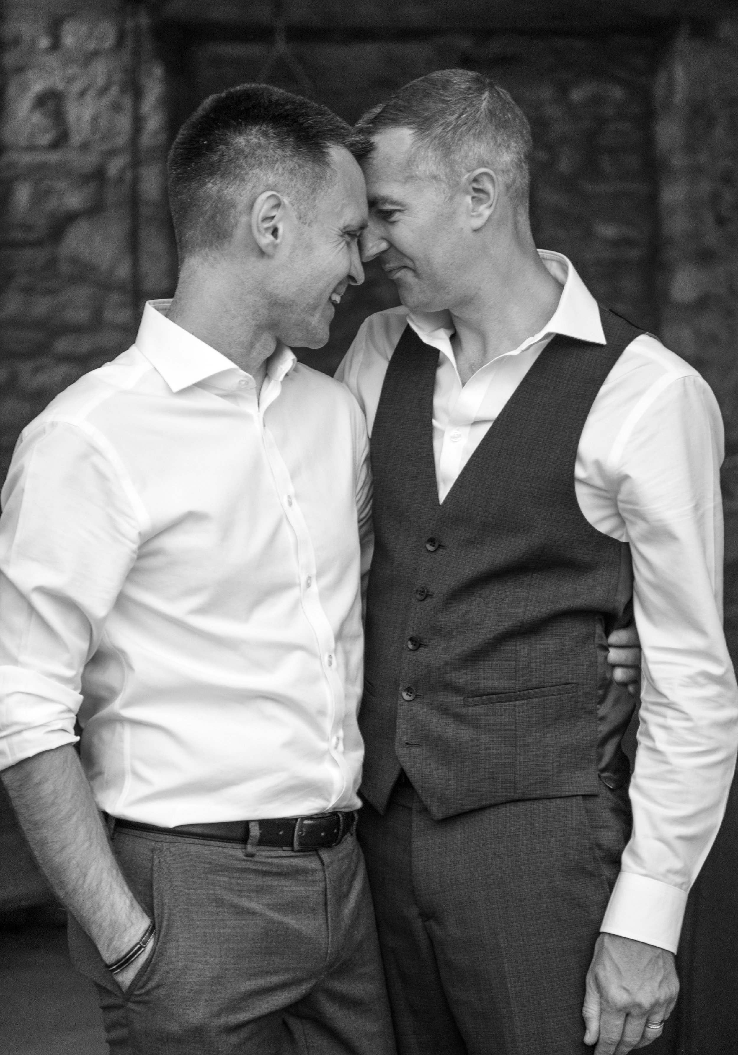 Two men smiling and touching foreheads, dressed in formal attire, in front of a brick wall.