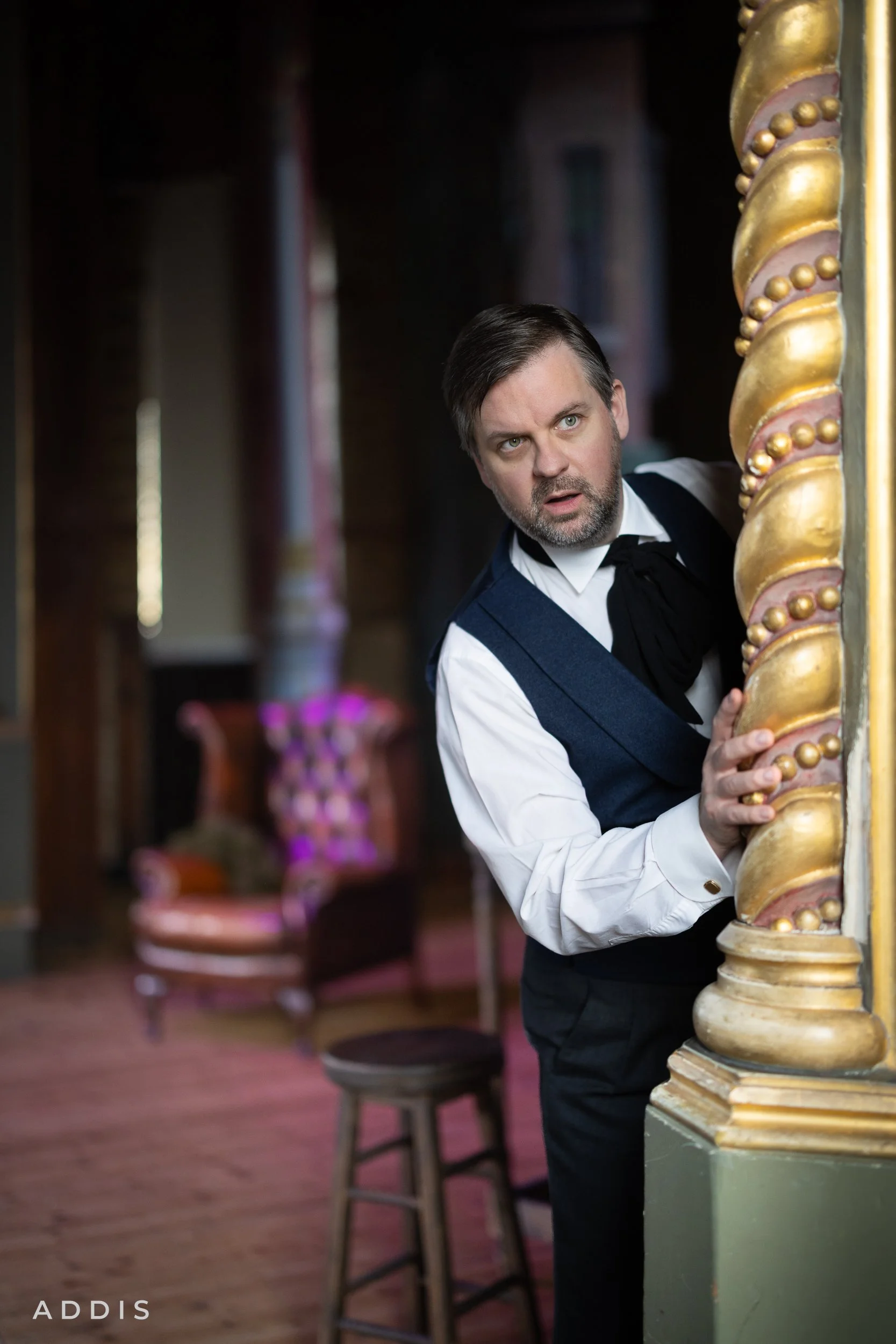 A man with a beard and dark hair peeks out from behind a decorated column in a room with antique furniture.