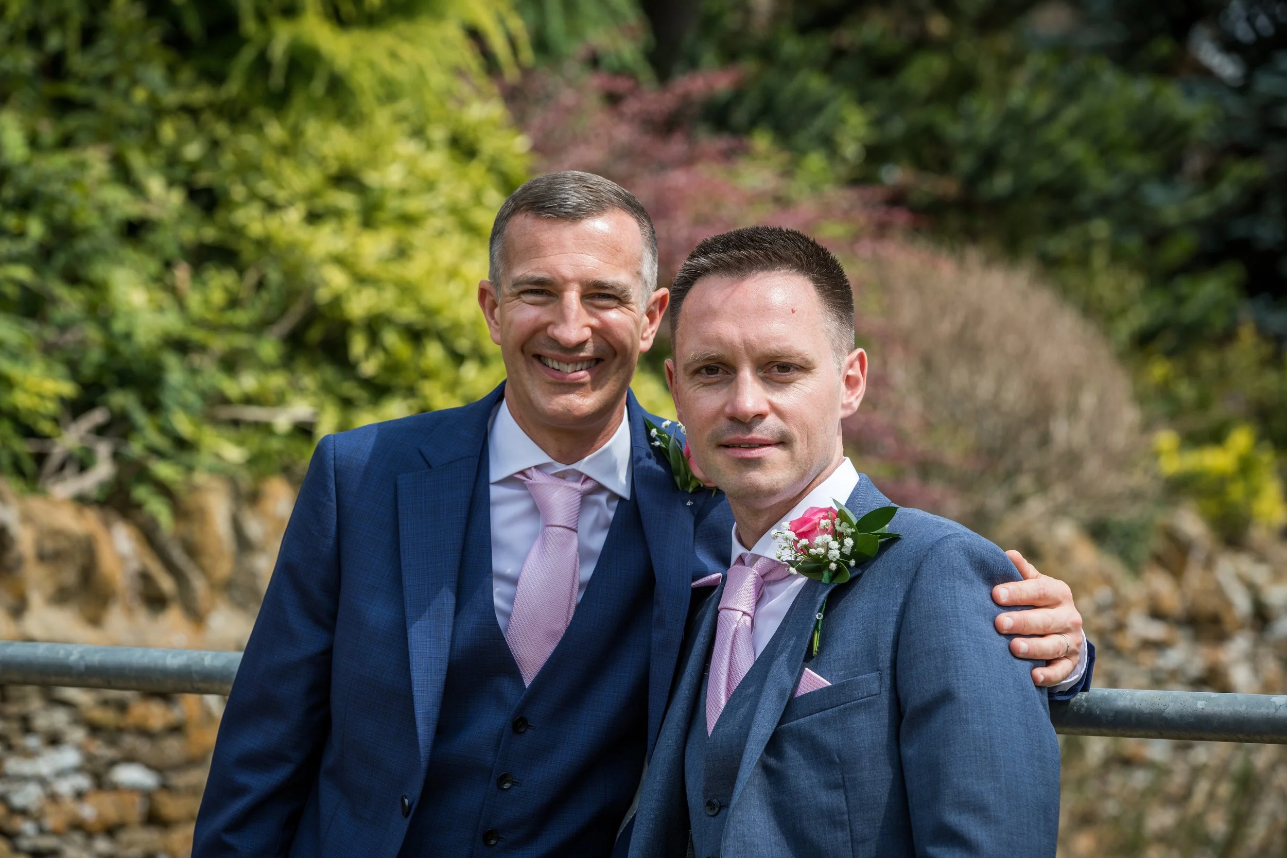 Two men dressed in blue suits with pink ties and boutonnières posing outdoors with greenery in the background.