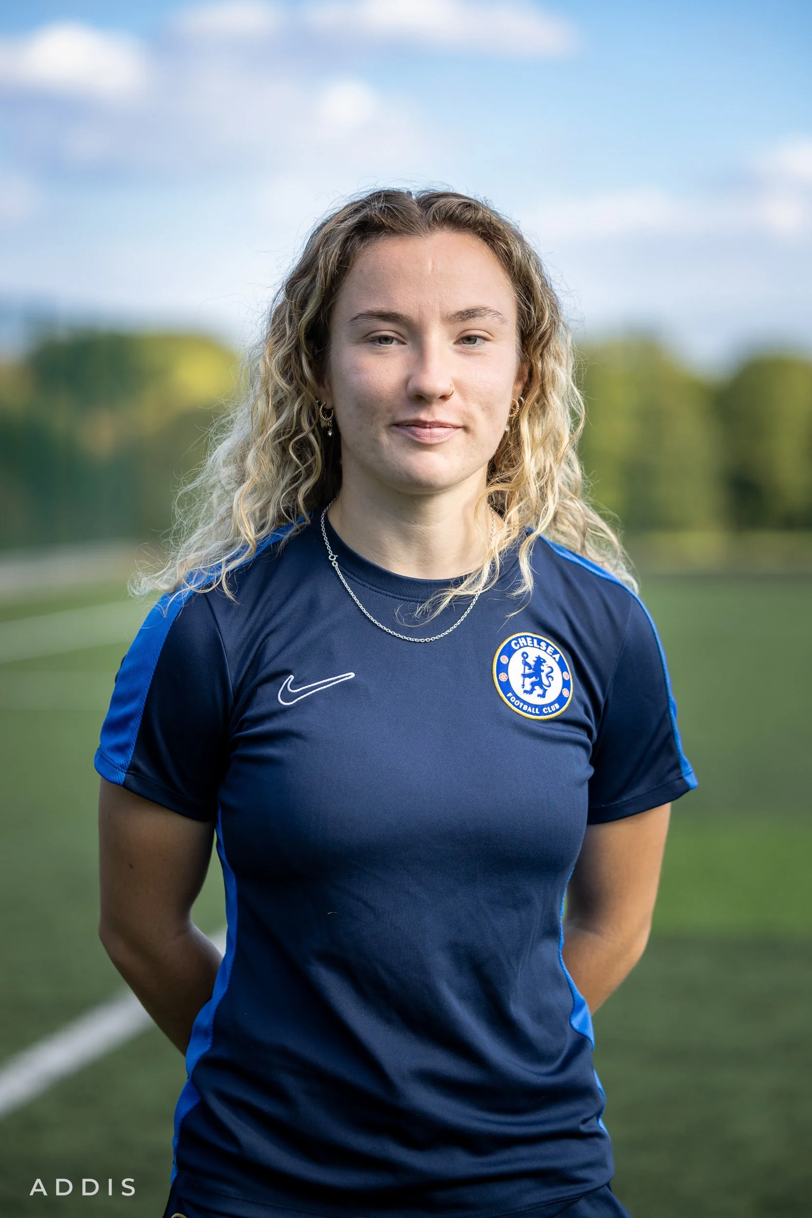 A young woman with long, blonde curly hair standing on a soccer field, wearing a Chelsea Football Club jersey, with a slight smile and outdoors background with trees and blue sky.