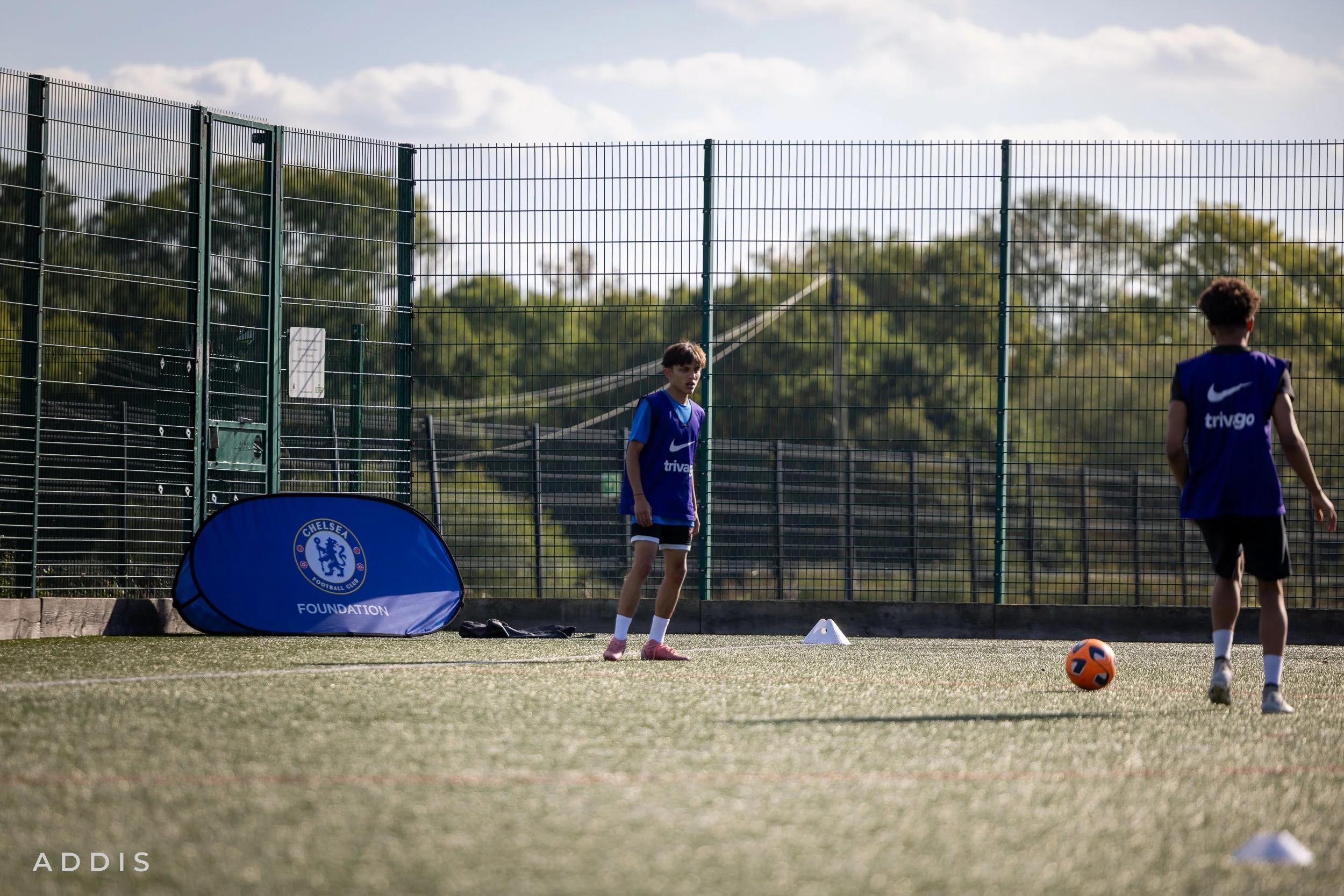 Two young boys in blue training vests practicing soccer on a field with orange and black ball, next to Chelsea FC Foundation sign, on a sunny day with a green fence and trees in the background.