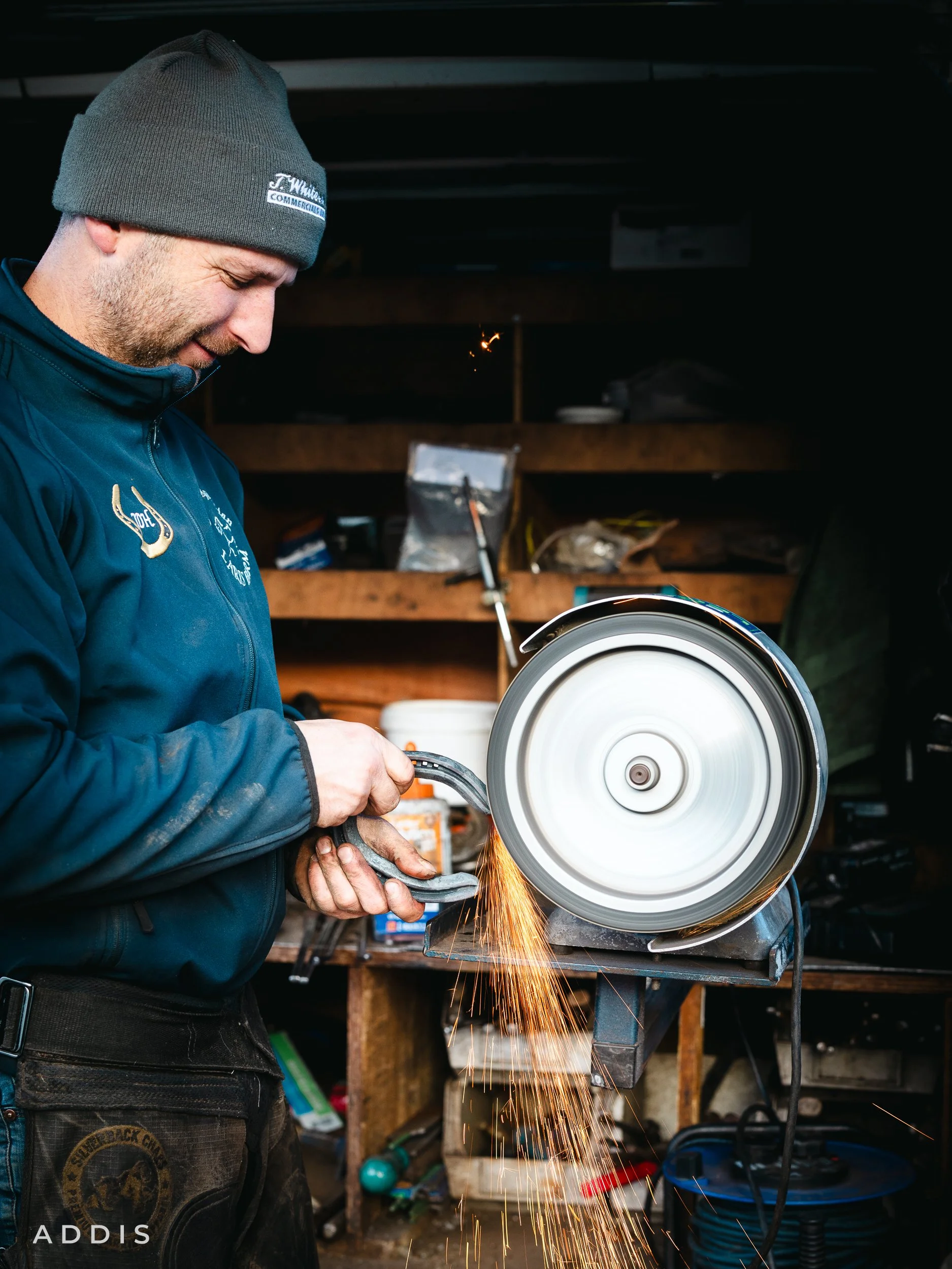 A man in a gray beanie and dark blue jacket is using a circular saw in a workshop, cutting a piece of metal, with sparks flying from the cut.