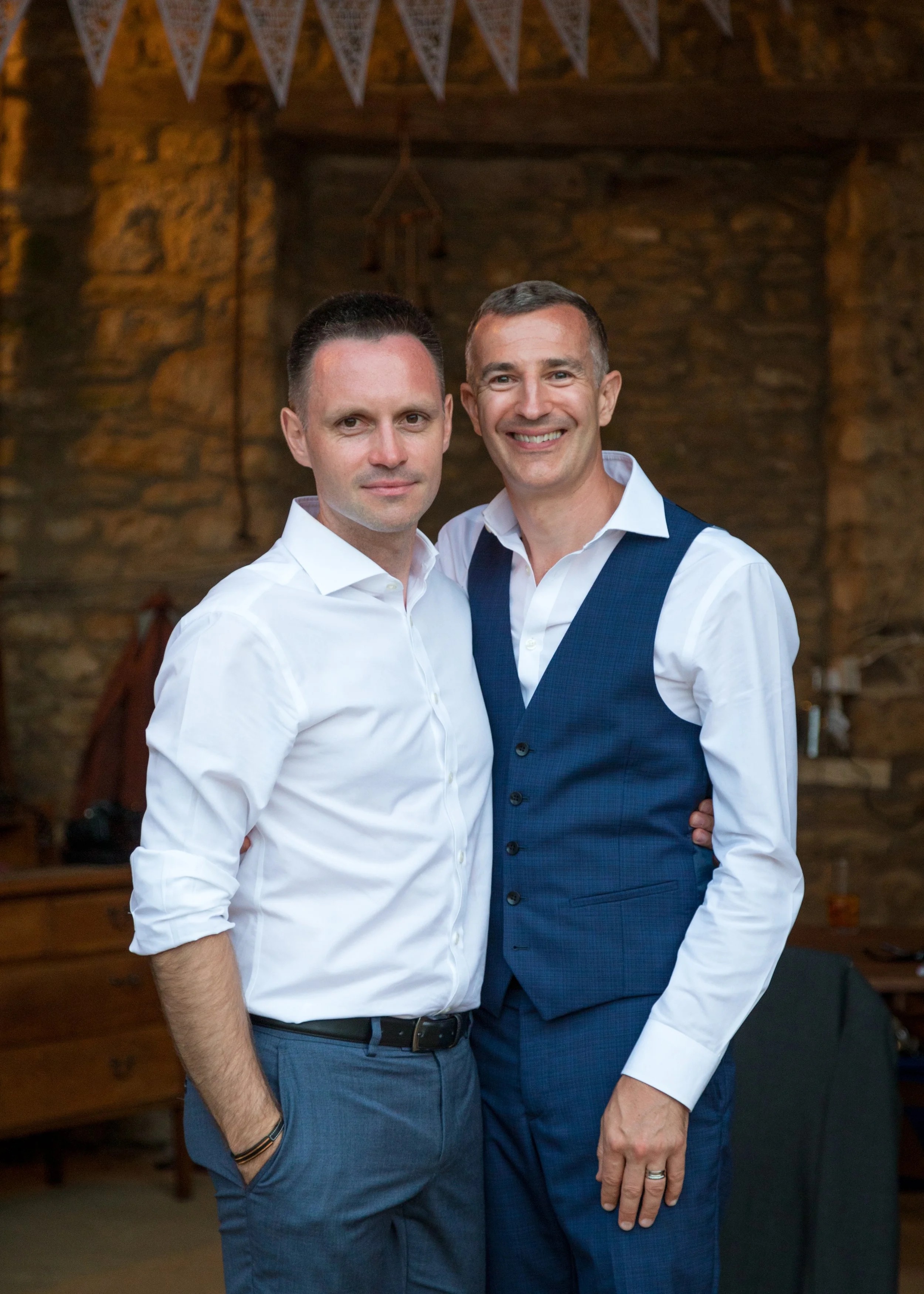 Two men standing inside a rustic wooden room, one with dark hair wearing a white shirt, the other with short hair wearing a blue vest and white shirt, smiling at the camera.