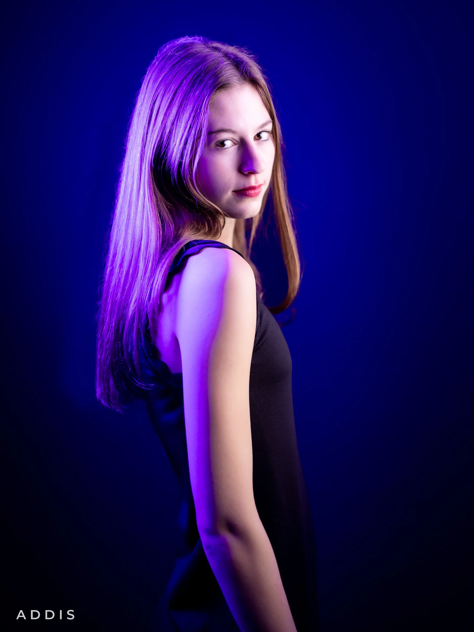 A young woman with long, straight brown hair and light skin poses against a black background illuminated with purple and blue lighting. She is wearing a sleeveless black dress and looks confidently at the camera.
