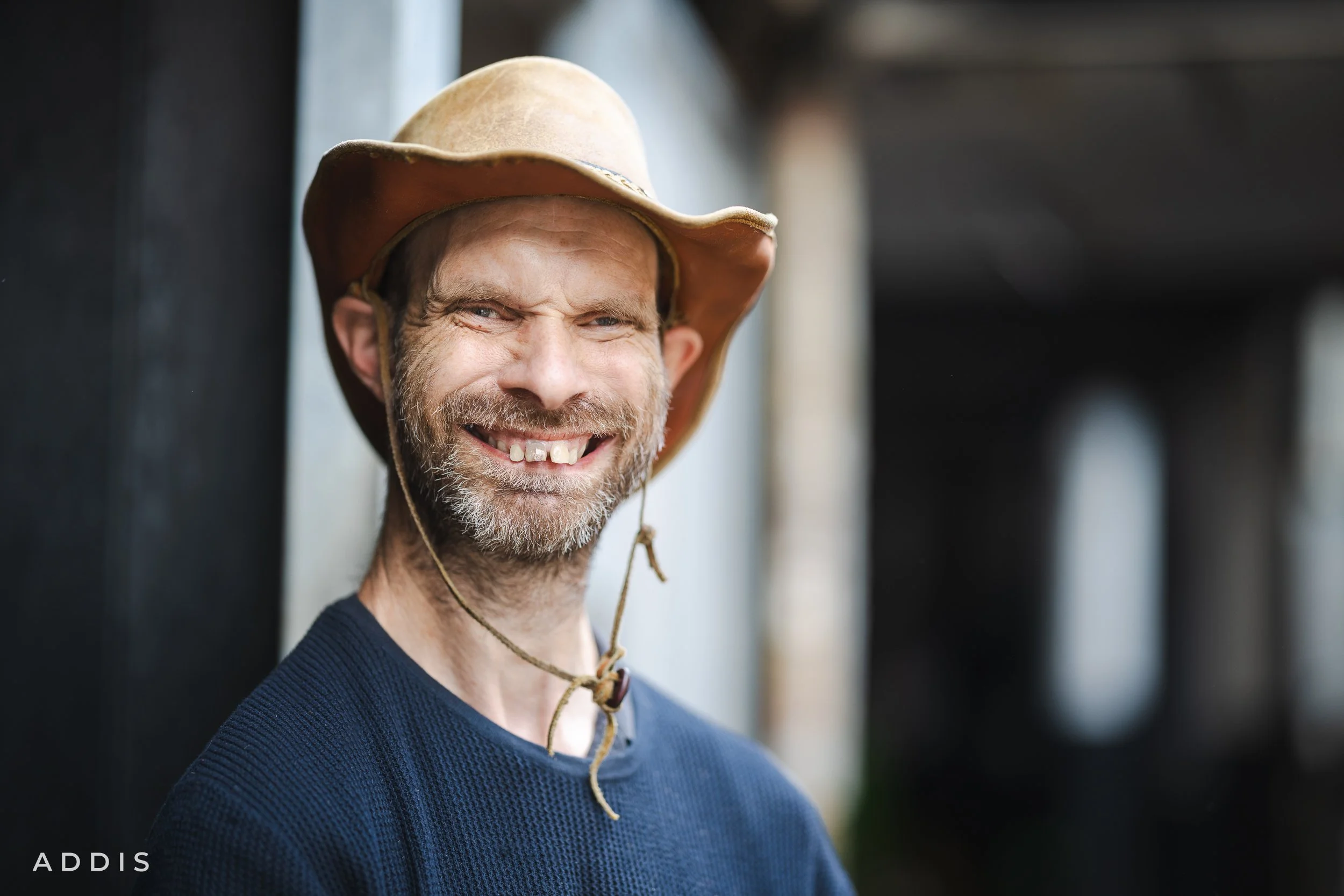 A smiling man with a beard wearing a cowboy hat and a navy blue sweater, standing outdoors near a black post.
