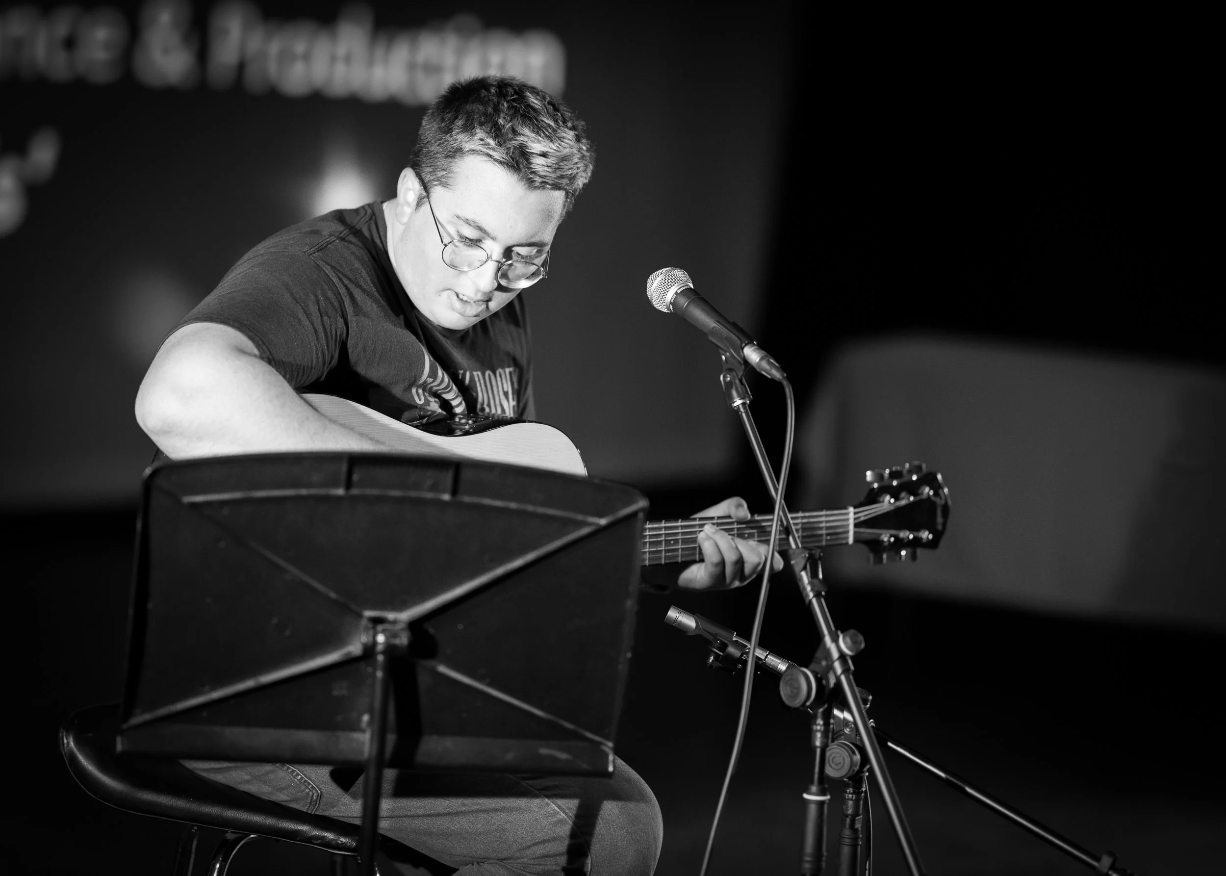 A young man with glasses playing an acoustic guitar on stage with a microphone in front of him, black and white photograph.