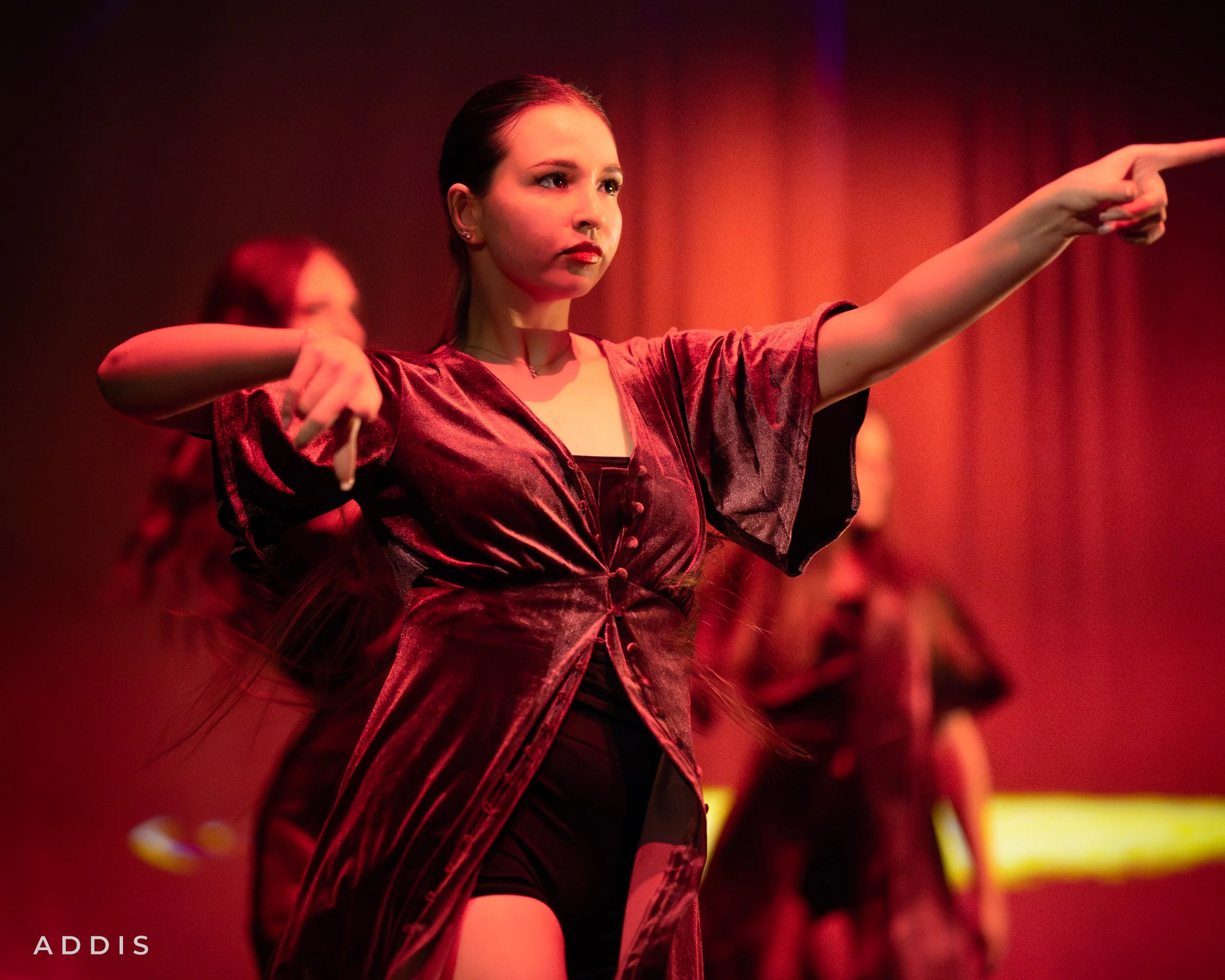 A young woman in a red velvet dress performing a dance move on stage with red lighting, with other dancers in the background.