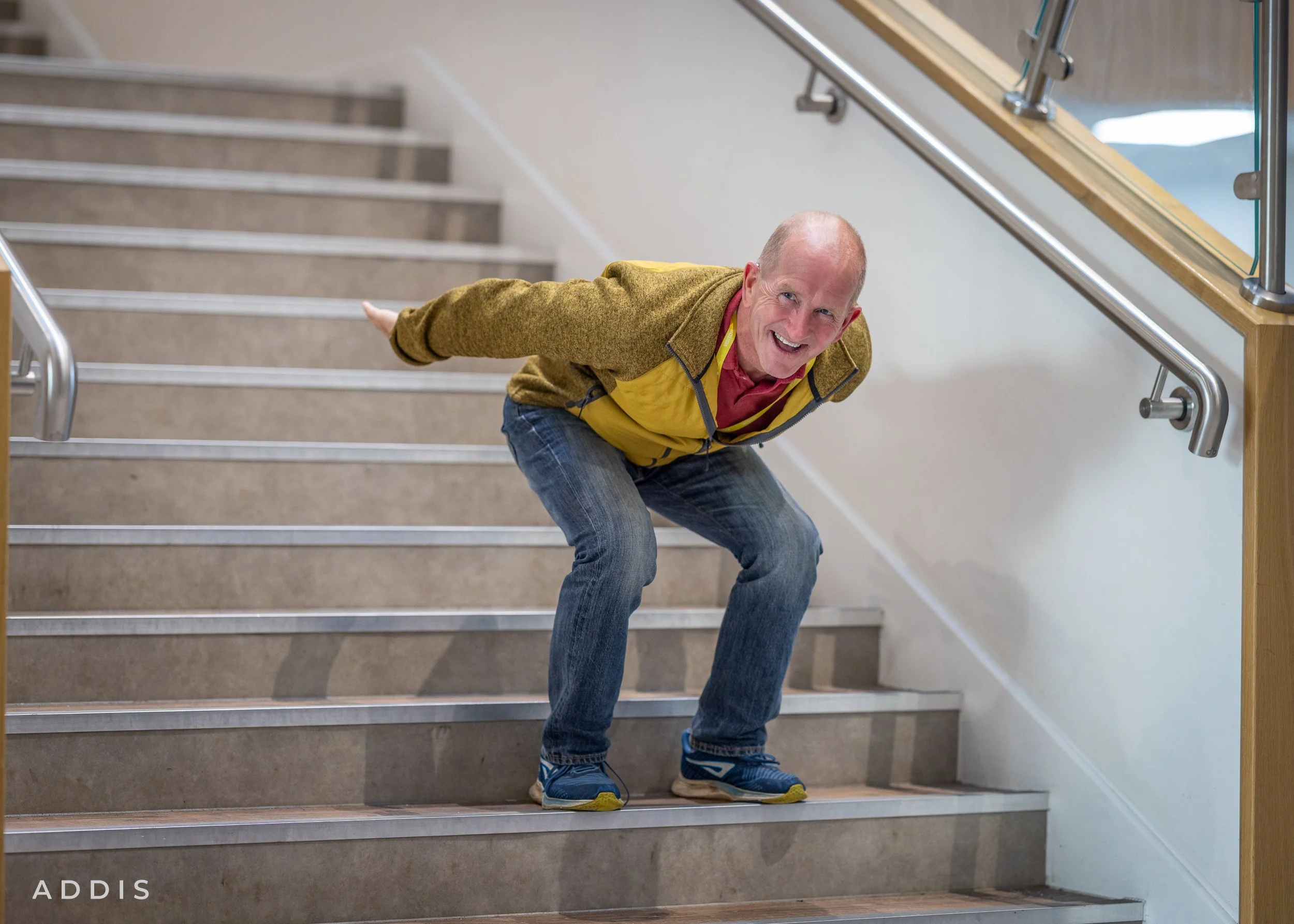 Eddie the Eagle Edwards smiling while bending forward on a staircase, wearing a brown jacket, yellow safety vest, blue jeans, and blue sneakers with yellow soles, holding onto the staircase railing.