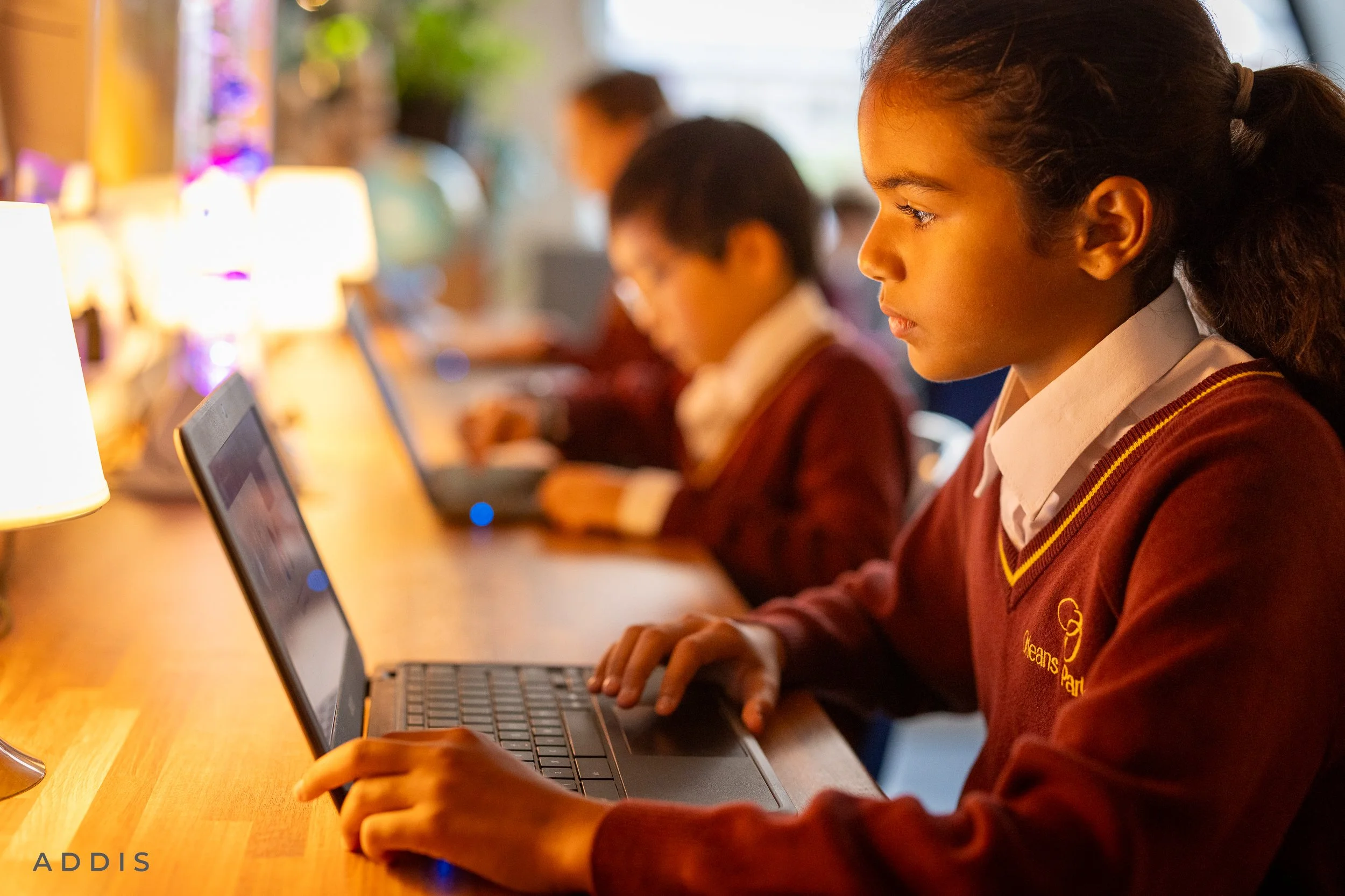 A young girl with curly hair tied back, wearing a school uniform and maroon sweater, sitting at a table using a laptop. In the background, other children are also engaged with devices, in a classroom or library setting.