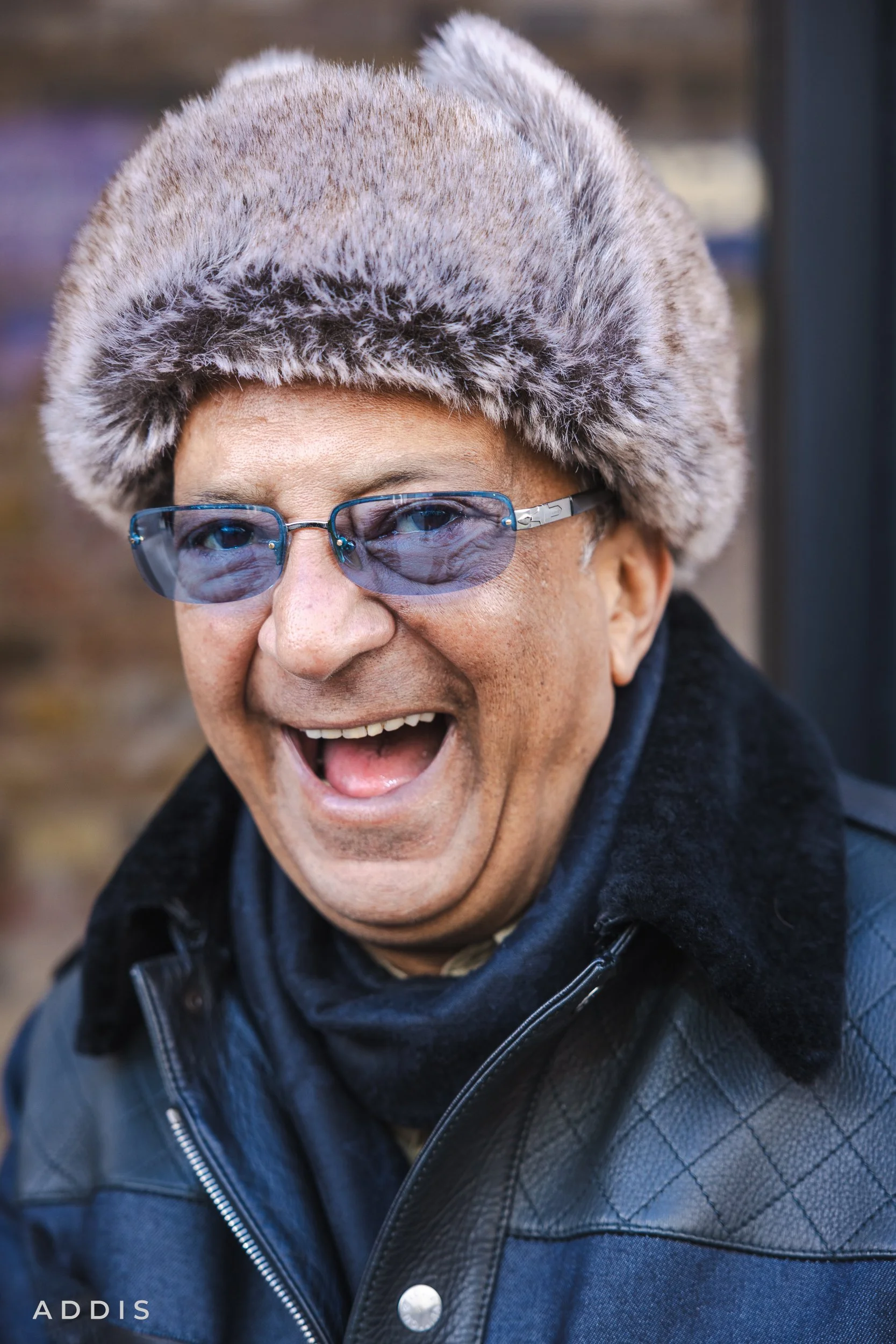 A smiling man wearing a furry winter hat and blue glasses, dressed in a black leather jacket and a black scarf.