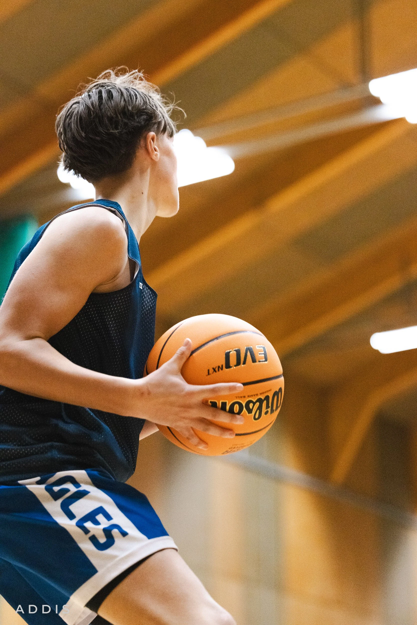 A female basketball player in a dark jersey holding a basketball, taken from a side angle inside a gym with wooden ceiling and fluorescent lights.