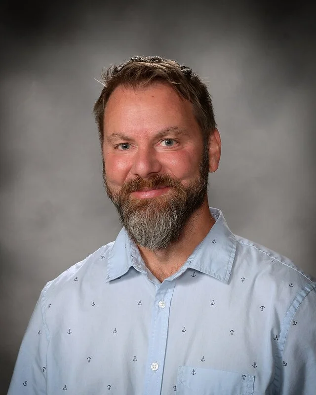 Portrait of a smiling man with light brown hair, a beard, and a mustache, wearing a light blue button-down shirt with small anchor patterns, against a gray background.