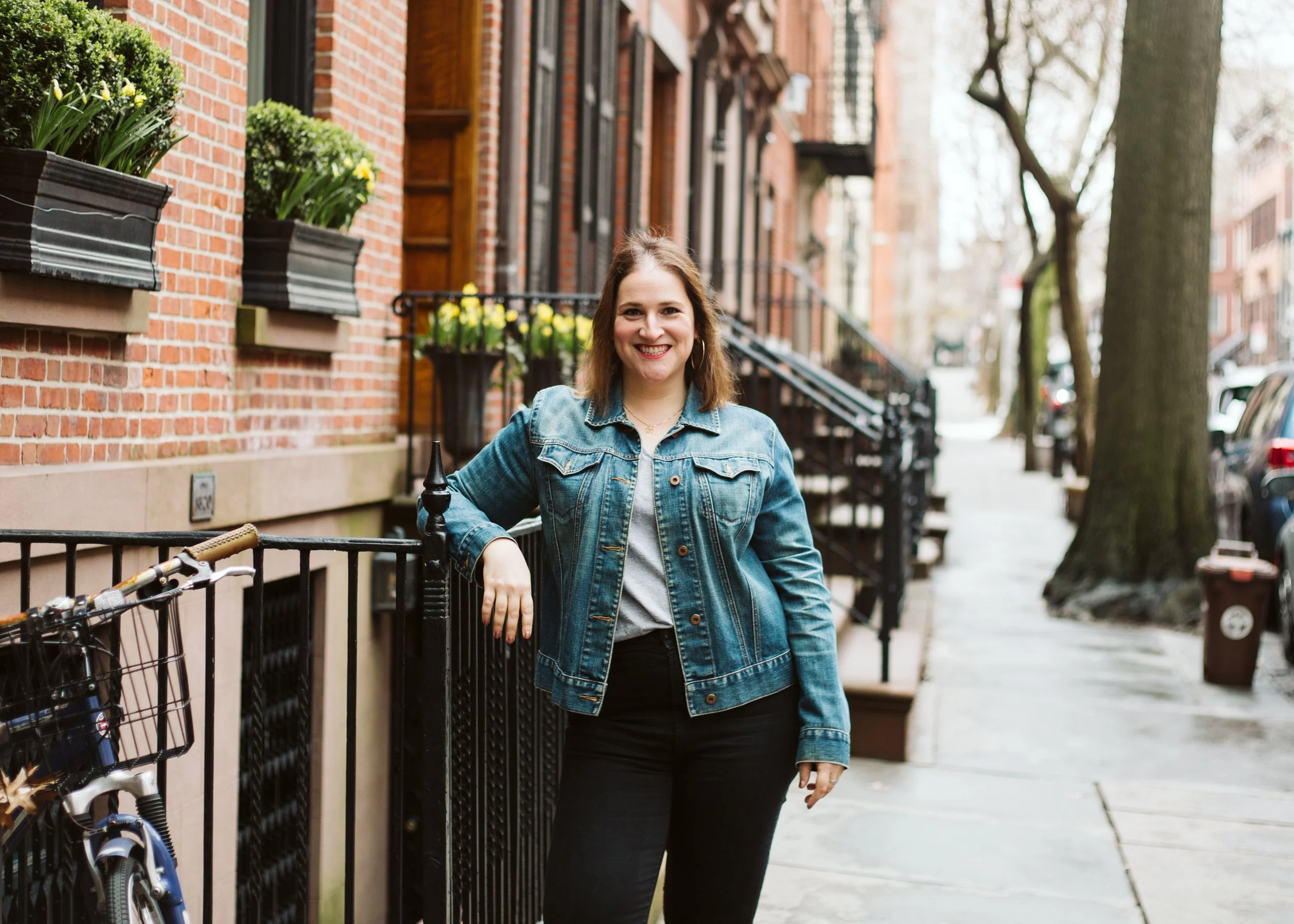 A woman with shoulder-length brown hair, wearing a denim jacket, smiling, standing on a city sidewalk next to a black metal railing. There is a bicycle with a basket parked against the railing. Behind her are brick residential buildings with flower boxes and trees lining the street.
