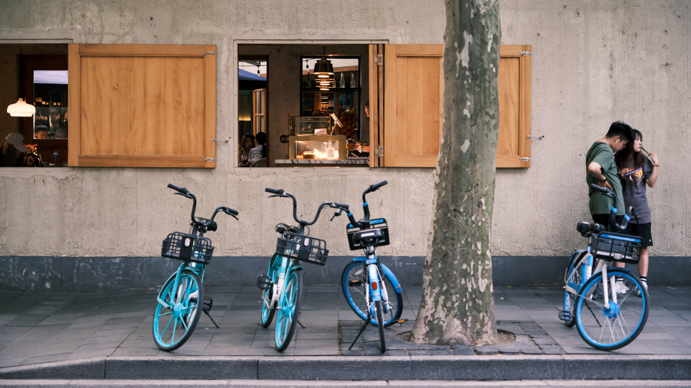 A beige stucco wall with open wooden window shutters, revealing a cafe interior with people inside. In front, four blue rental bikes are parked on the sidewalk, and a large tree trunk is visible. Two people stand near the bikes, looking at a phone.