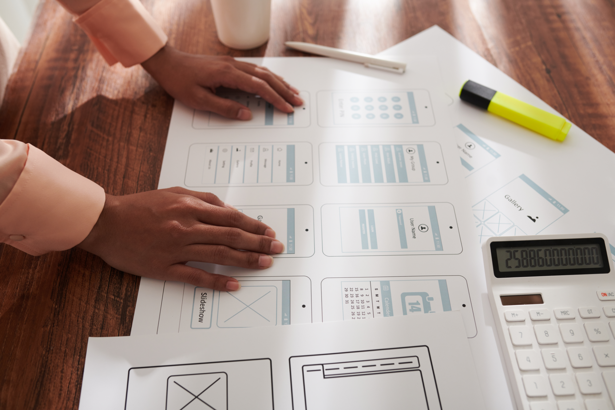 Person working on a wireframing or UX design project with mobile app sketches on printed paper, a white pen, yellow highlighter, calculator, and a white cup on a wooden table.