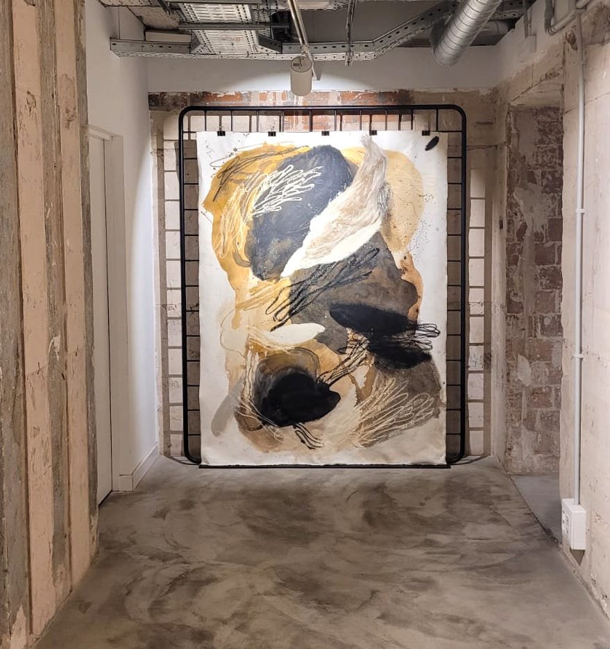 Pintura abstracta con colores marrón, negro, beige y blanco colgada en una pared de ladrillo en un pasillo industrial con paredes y techo expuestos.