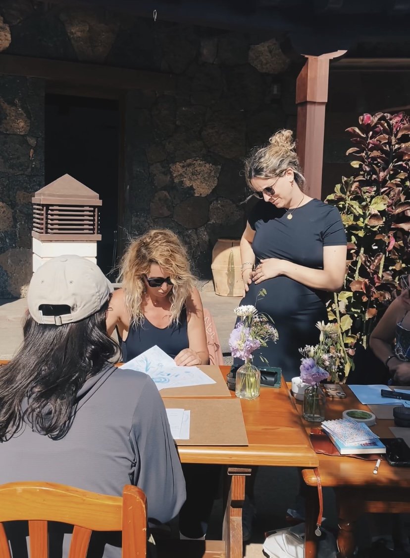 Un grupo de personas alrededor de una mesa al aire libre, una persona sentada revisando papeles y otras de pie observando. Hay flores en la mesa. El entorno parece ser un jardín.