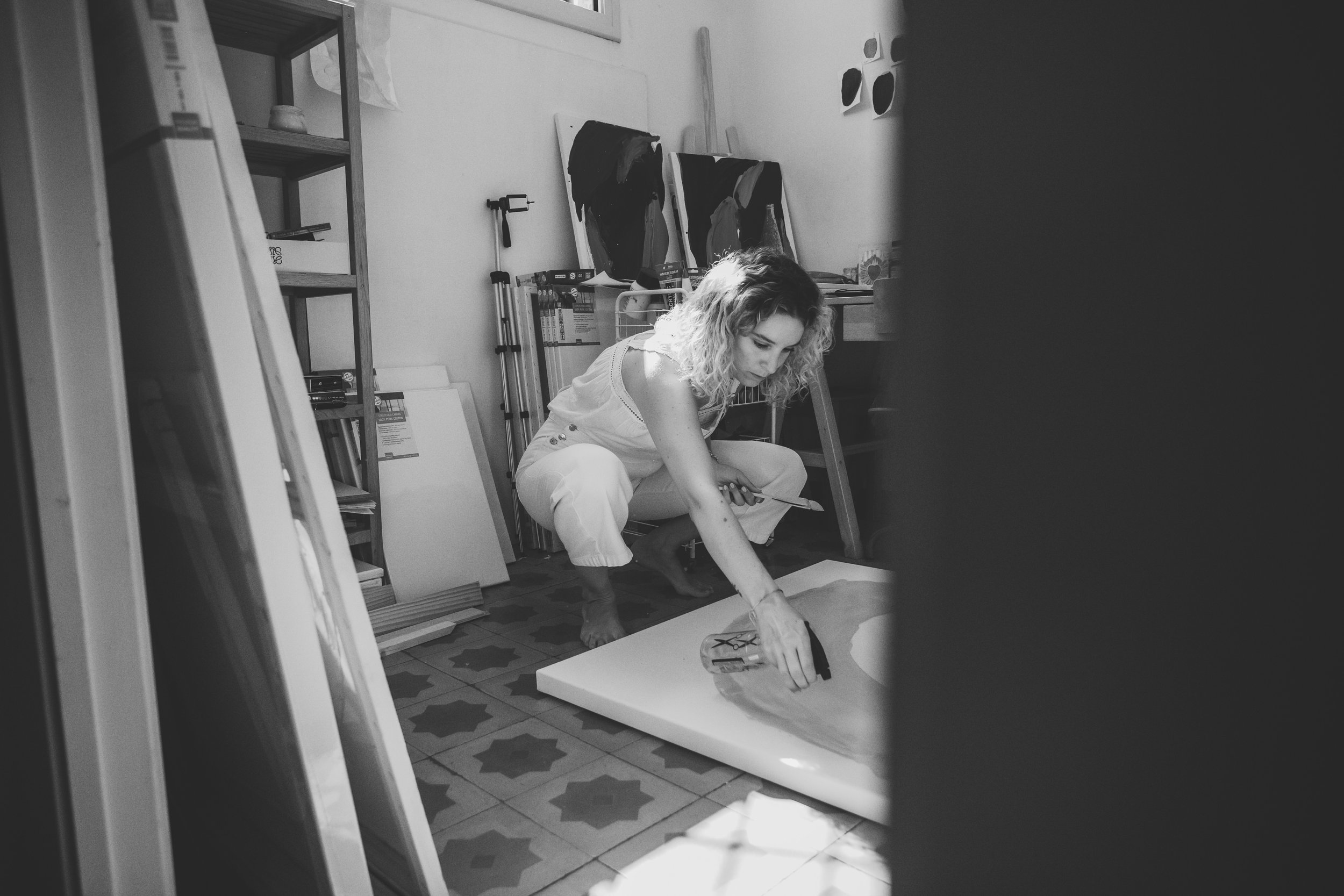 Mujer pintando sobre un lienzo en un estudio de arte, rodeada de materiales y obras inacabadas.