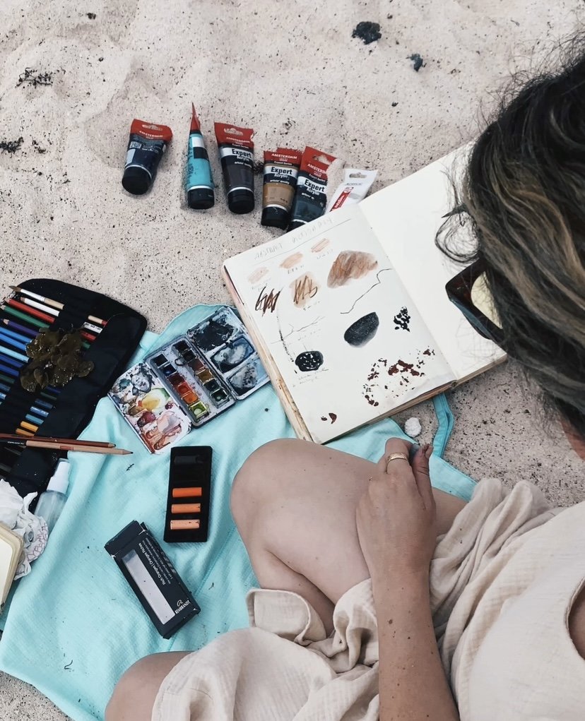Persona sentada en la arena con materiales de arte. Hay tubos de pintura acrílica, un estuche con lápices de colores, acuarelas, pasteles y un cuaderno de bocetos abierto con diseños. Se observa parte del brazo y pierna de la persona.