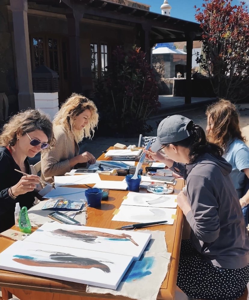 Grupo de personas pintando en una mesa al aire libre, con materiales de arte como acuarelas y pinceles.