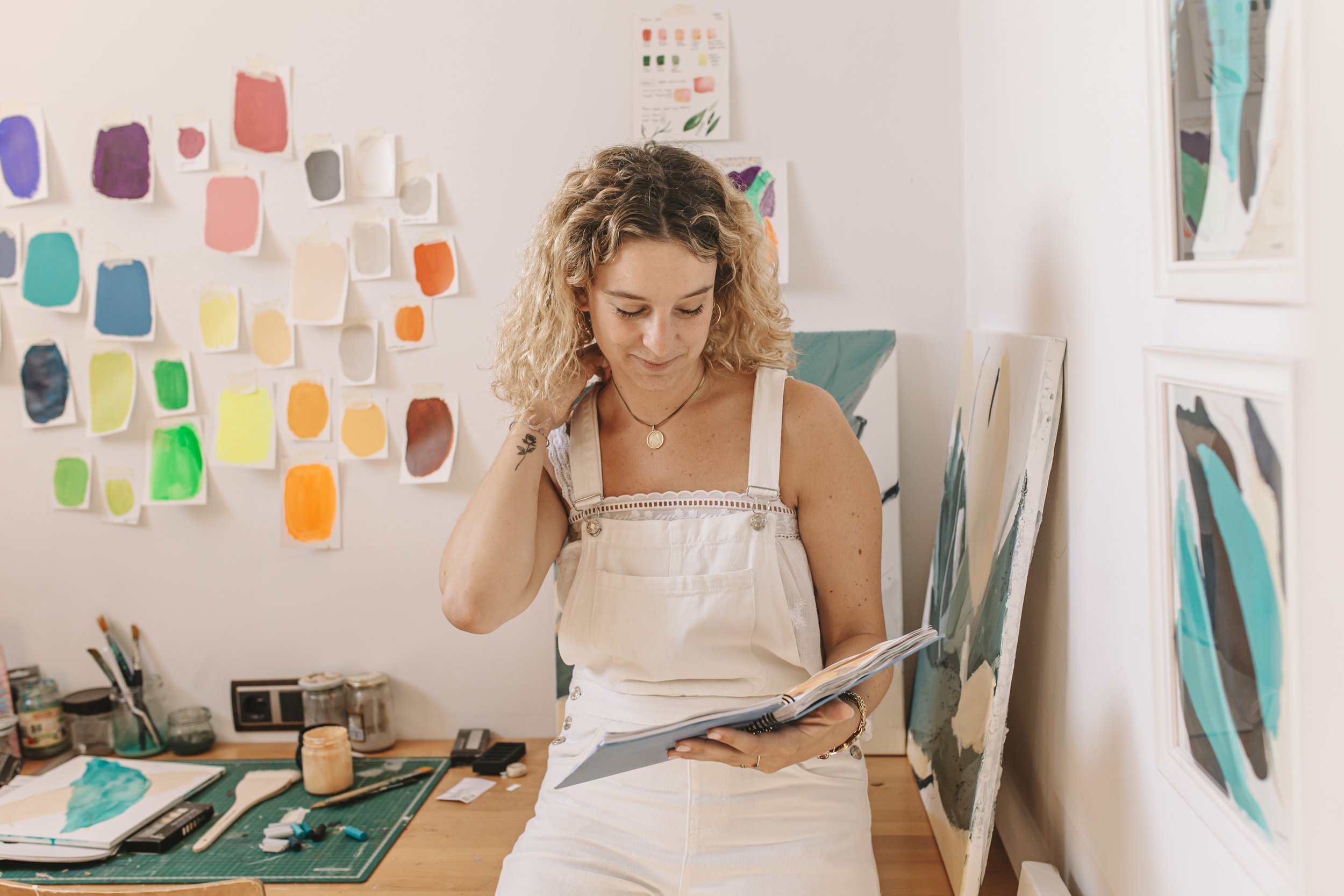 Mujer con pelo rizado sosteniendo un cuaderno en un estudio artístico, con muestras de color en la pared y material de arte en la mesa.