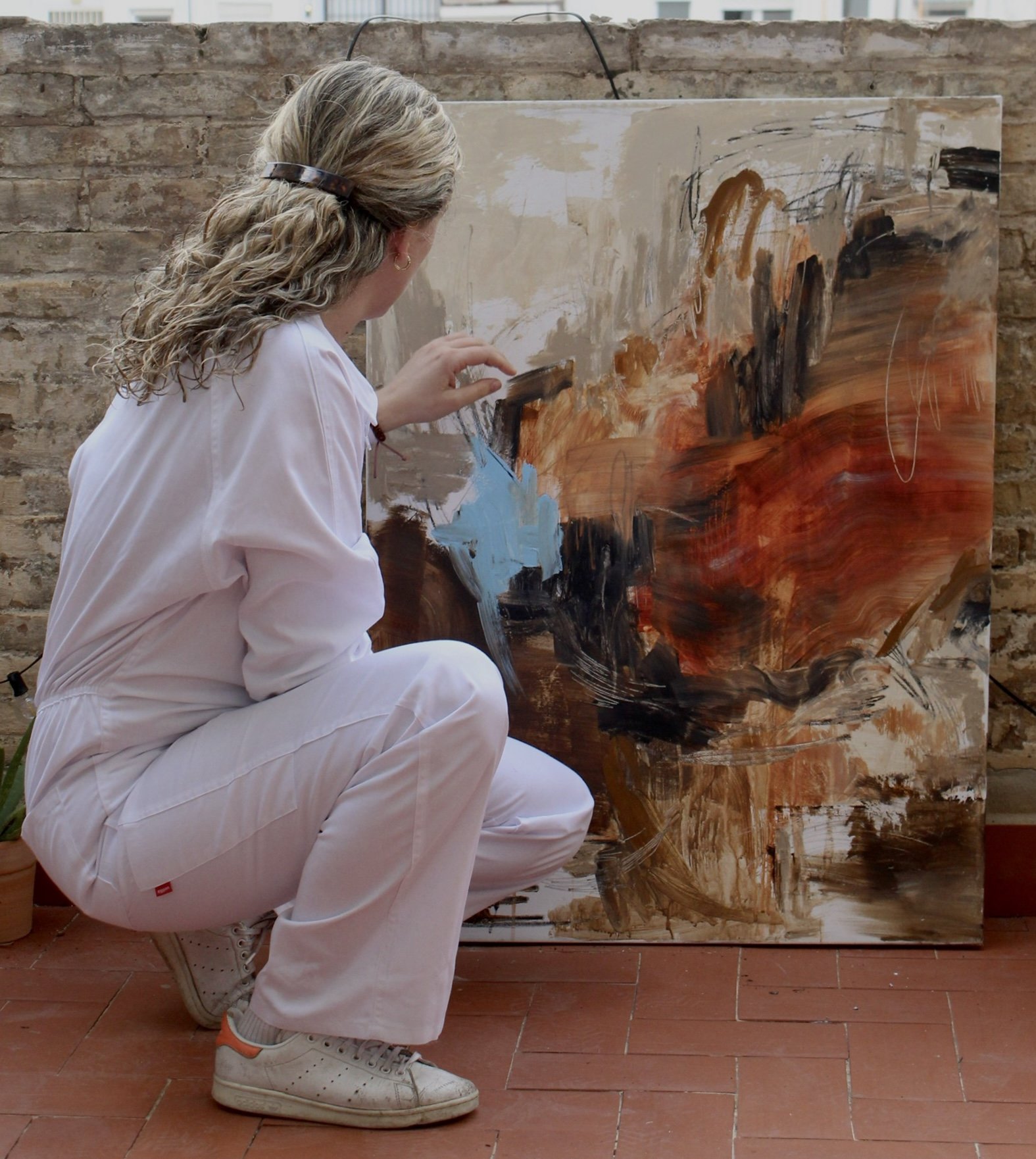 Mujer pintando un cuadro abstracto en un patio con paredes de ladrillo, vistiendo un mono blanco y zapatillas deportivas.