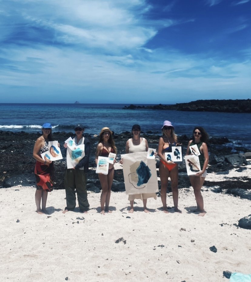 Grupo de personas en la playa mostrando pinturas frente al océano.