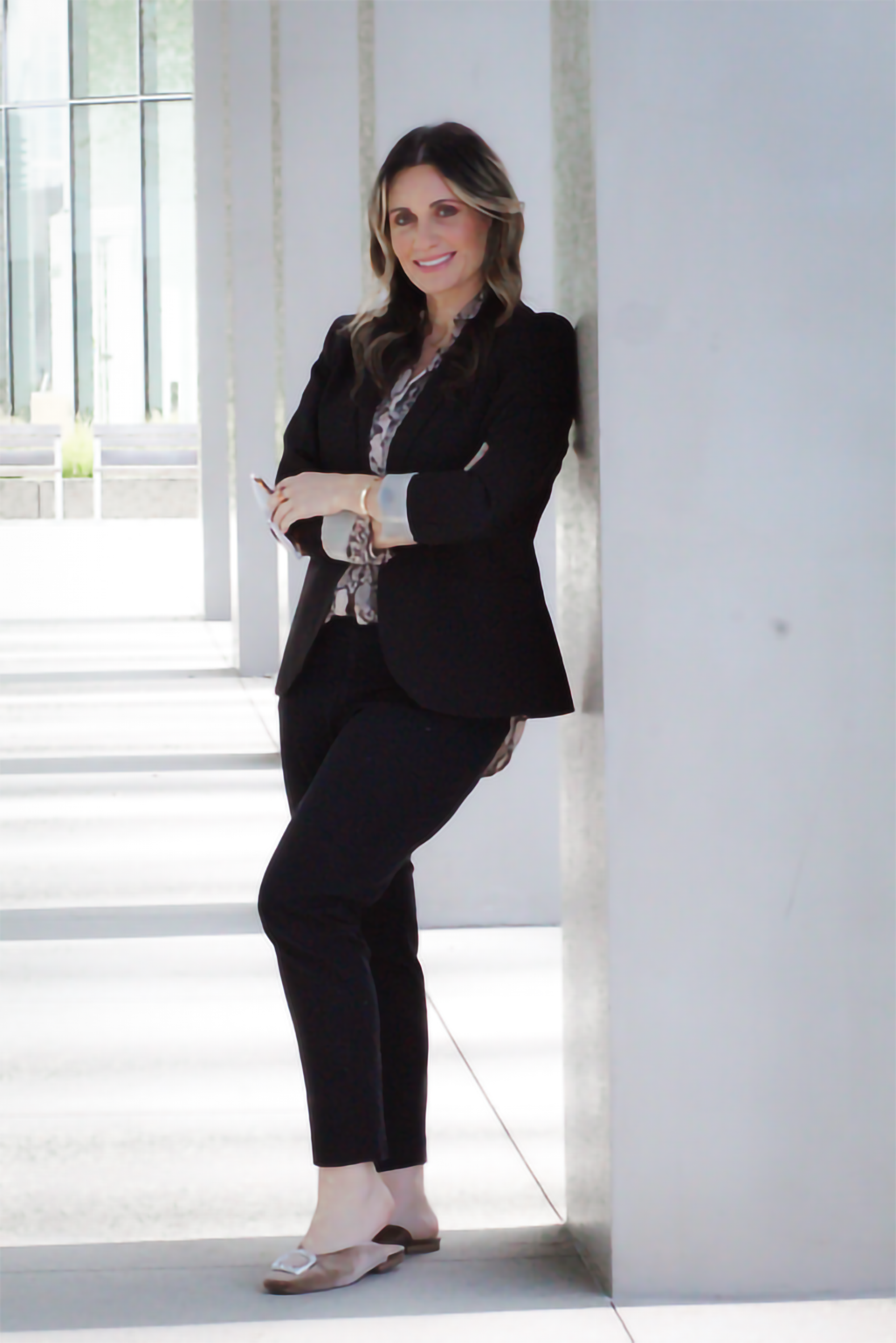 A woman in a black suit and patterned blouse leaning against a wall in a bright modern building, smiling with arms crossed.