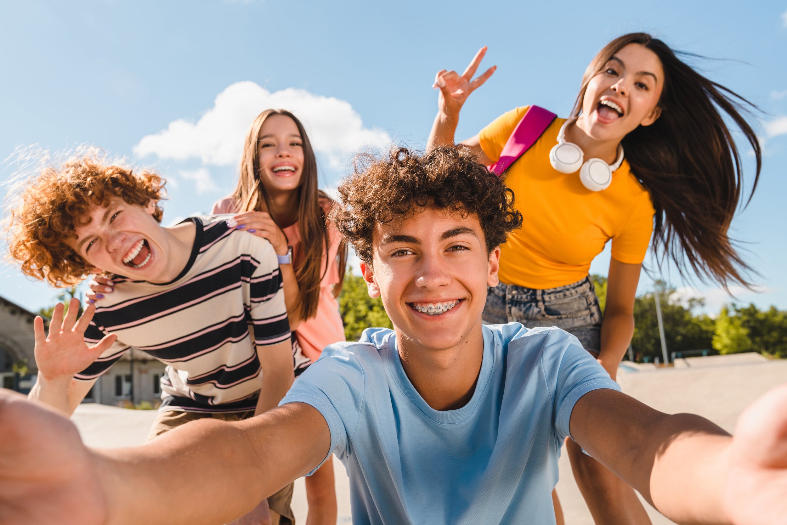 Four teenagers taking a selfie outdoors on a sunny day, smiling and waving, with blue sky and clouds in the background.